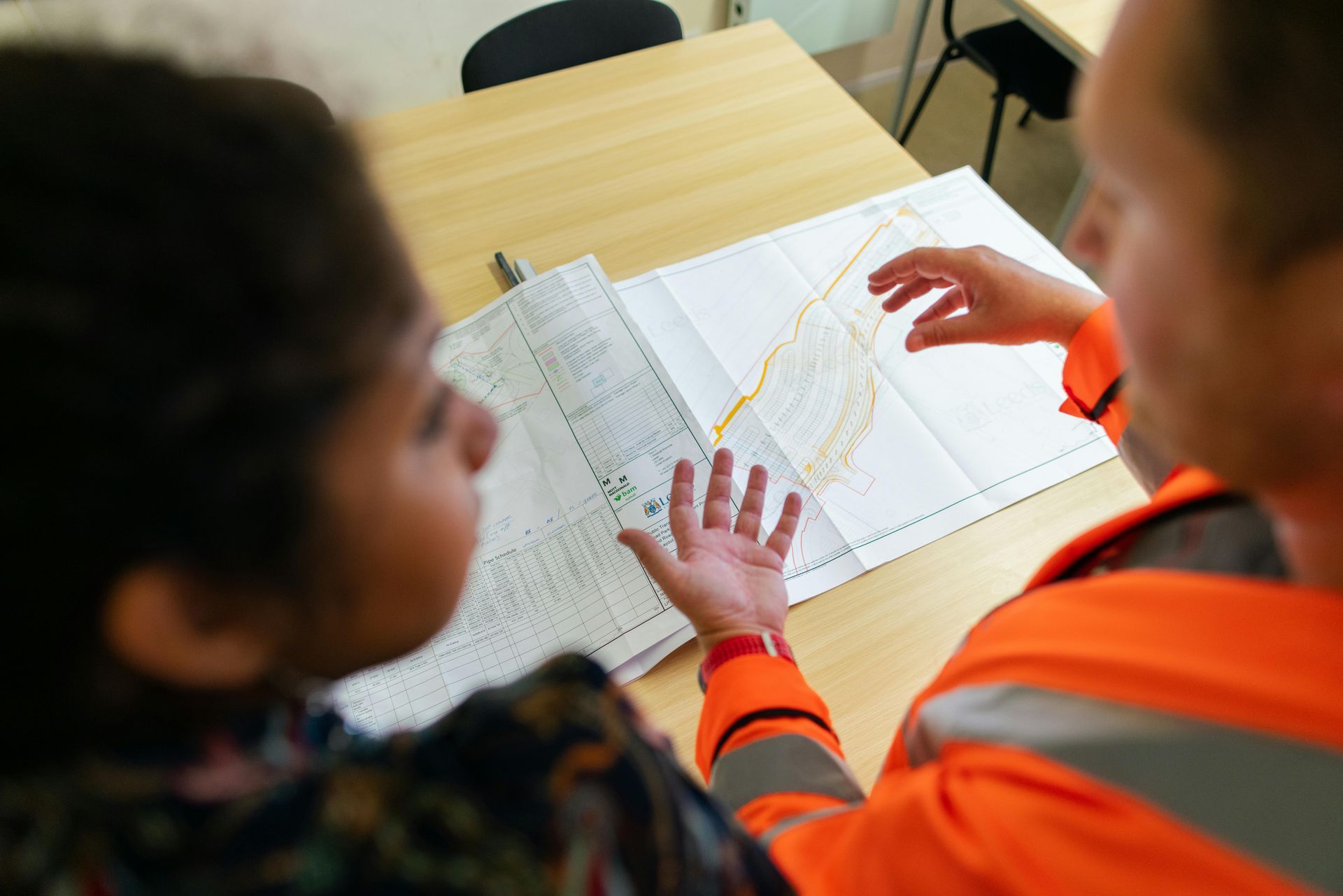 Two people in orange safety vests reviewing blueprints at a desk with a ruler and pencil