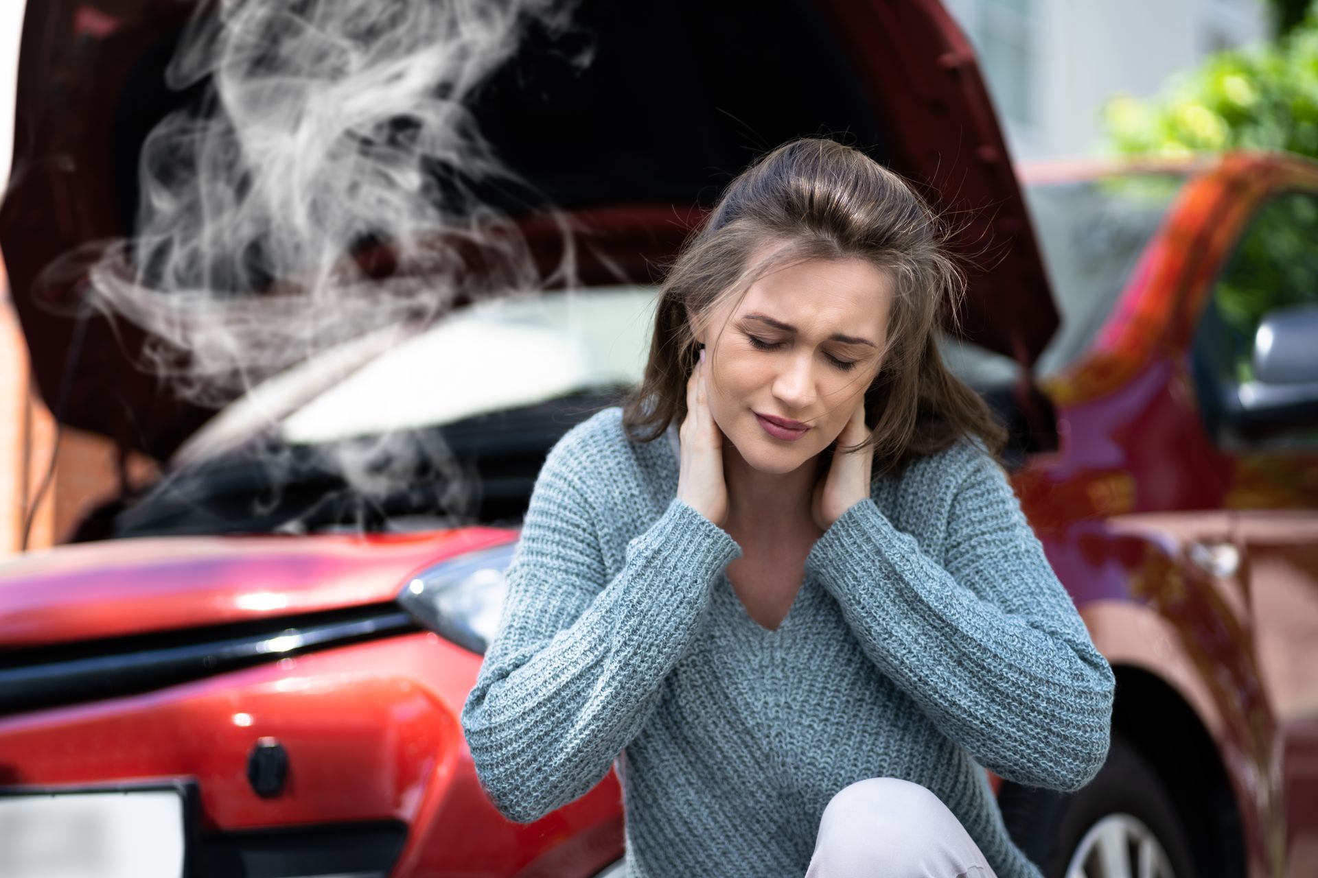 A woman is sitting in front of a broken down car with smoke coming out of the hood.