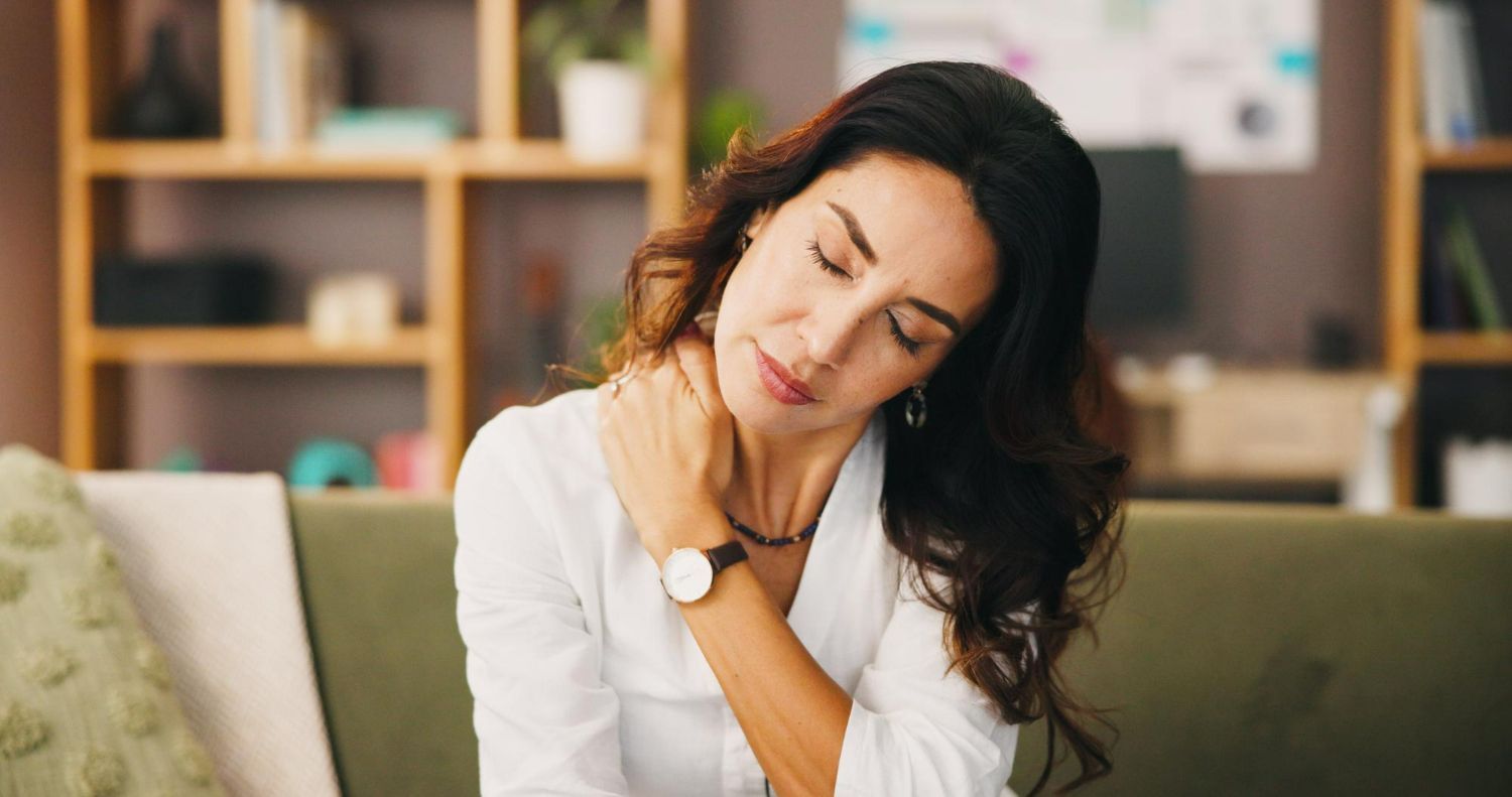 A woman in a white shirt sits on a sofa, eyes closed, holding her neck in discomfort.