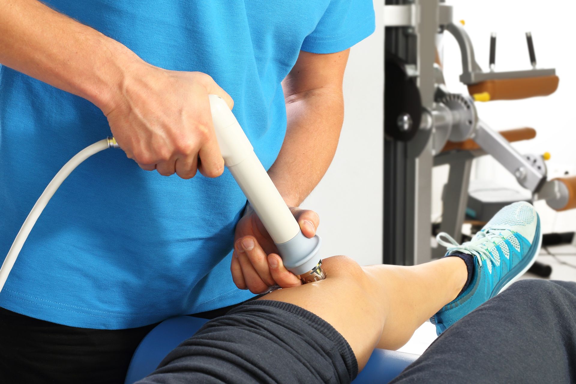 Therapist using a medical device on a patient's knee. Blue shirt, gym setting.