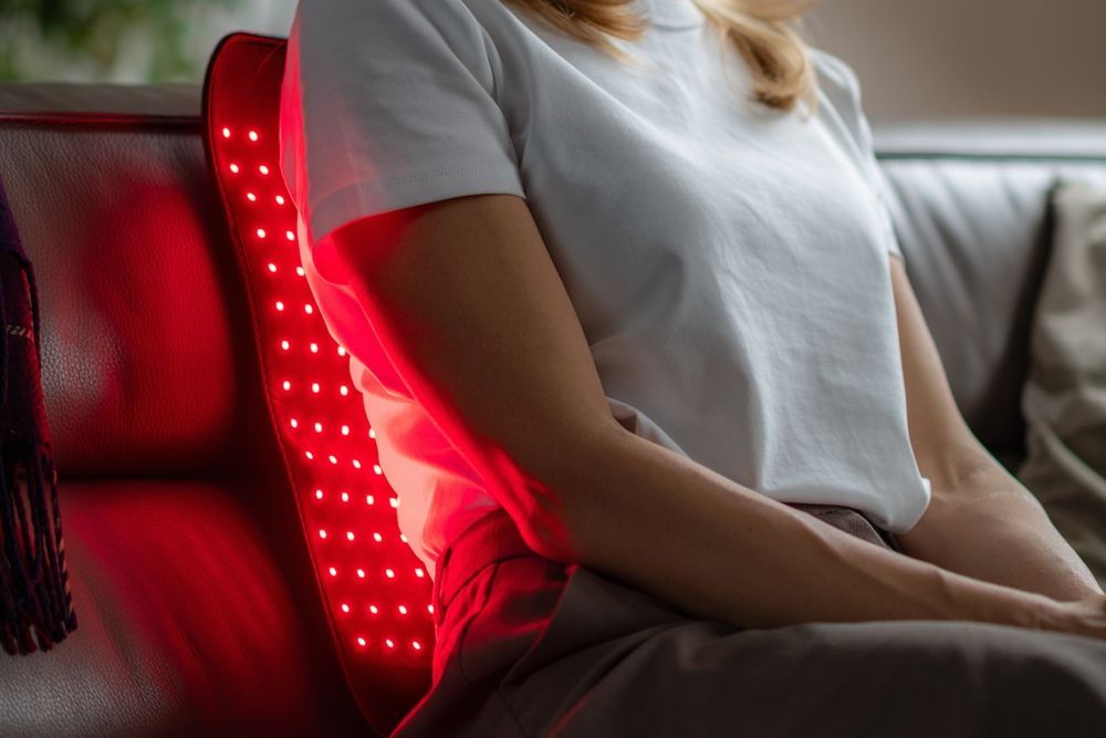 A person sits on a sofa using a red light therapy panel placed behind their back.