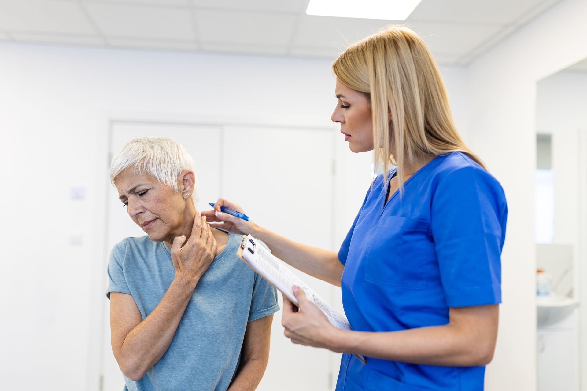 Nurse examining an older person's neck in a white medical setting. The person holds their neck, showing discomfort.