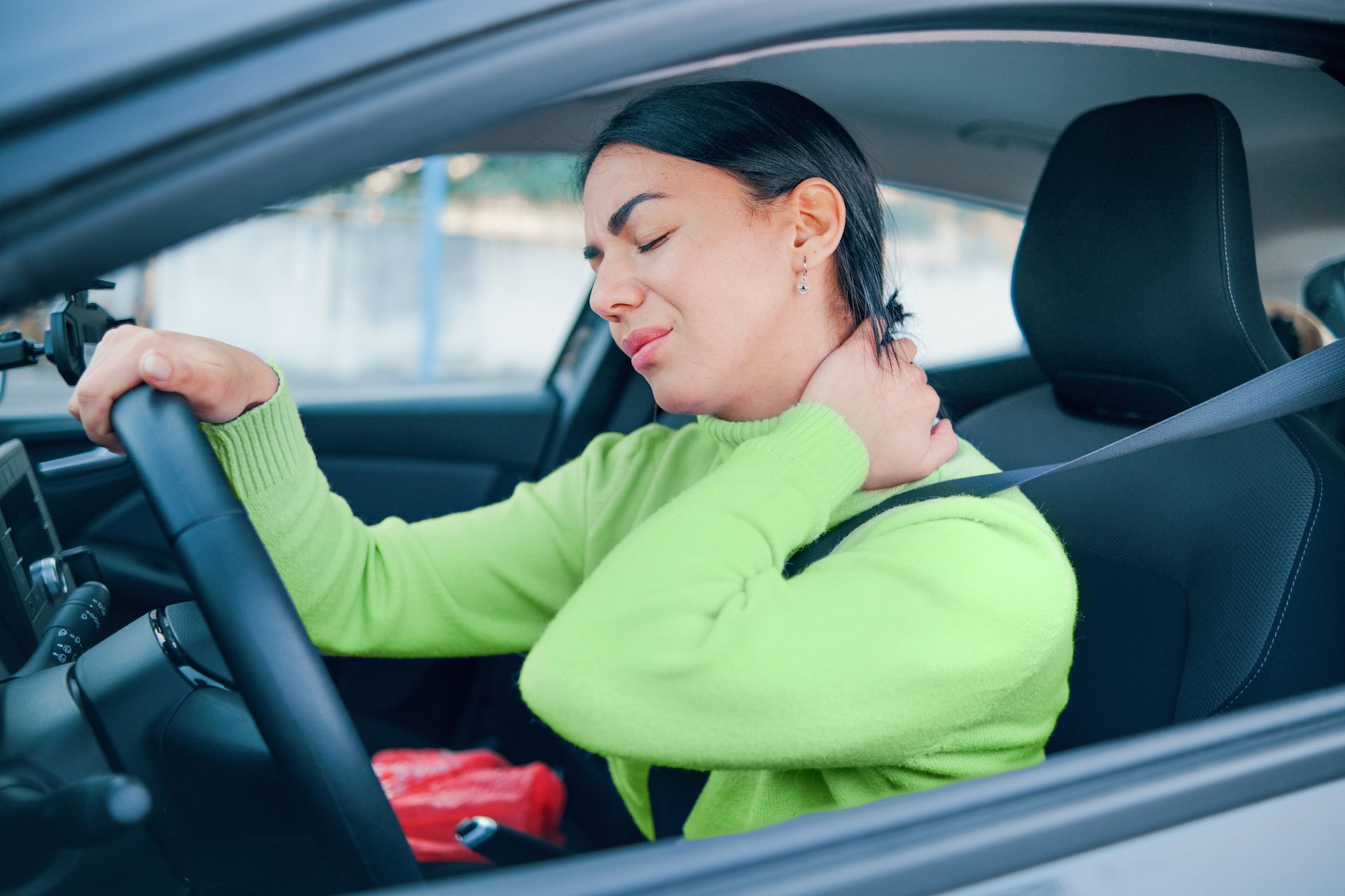 Woman in car, grimacing while holding her neck, likely experiencing neck pain.