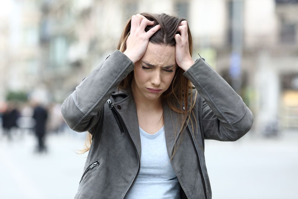 A person in a gray jacket with a distressed expression, hands clutching their head, standing in a blurred city setting.