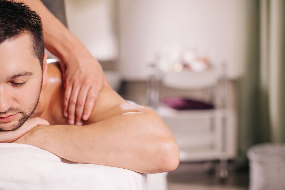 A person receives a shoulder massage while resting their head on their arms in a calm, dimly lit room.