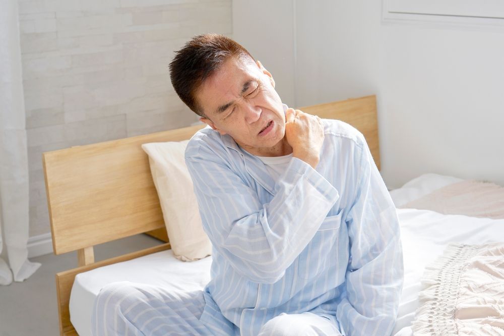 A person in light-colored pajamas sits on a bed, wincing and clutching their shoulder in pain.