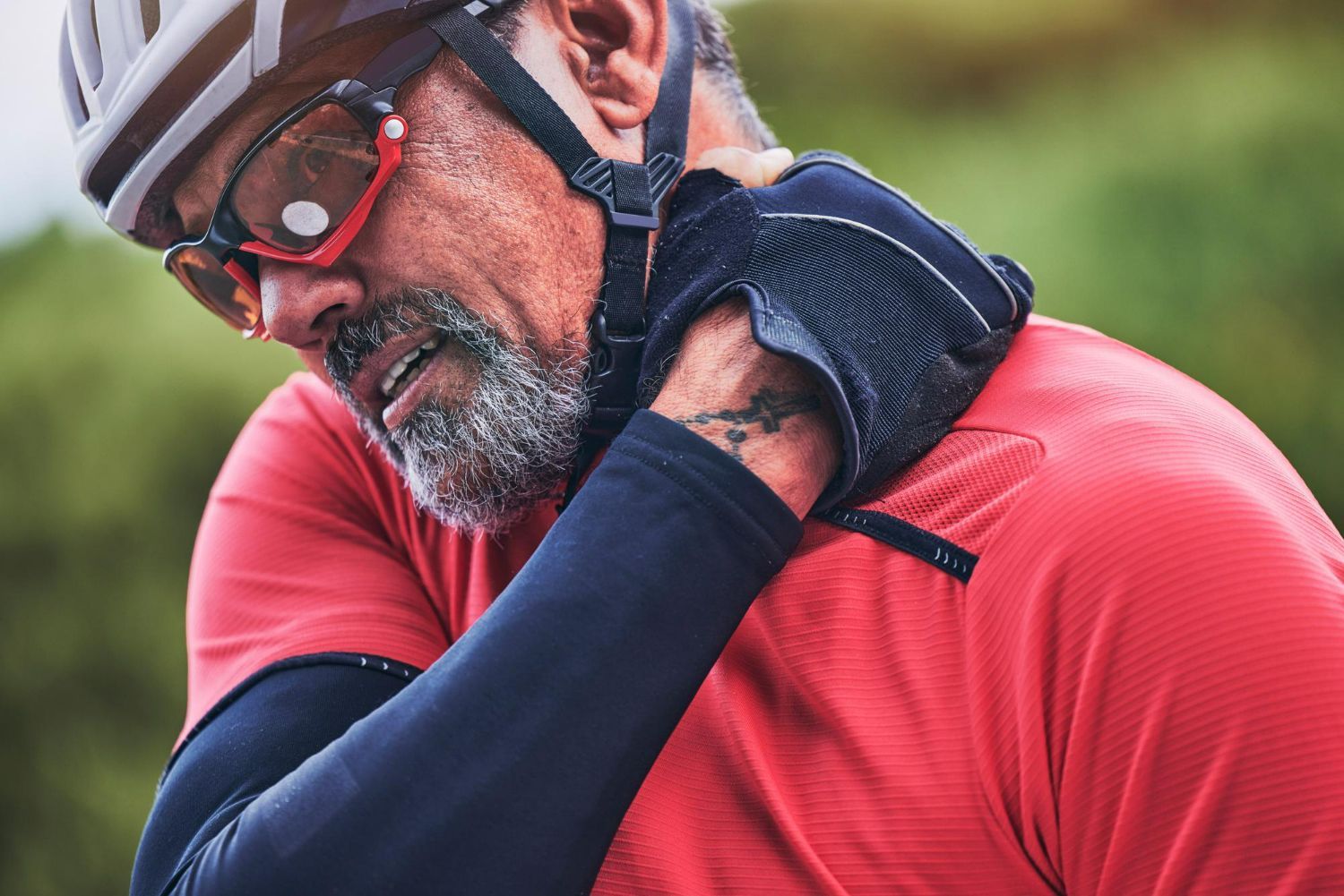 Cyclist clutching his neck, likely in pain. He wears a helmet, sunglasses, and a red shirt; outdoors.
