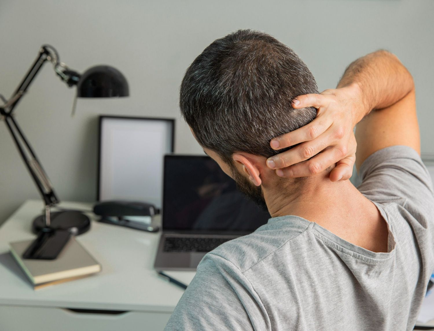 Man at desk, hand on neck, showing pain; laptop, desk lamp, phone, notebook.