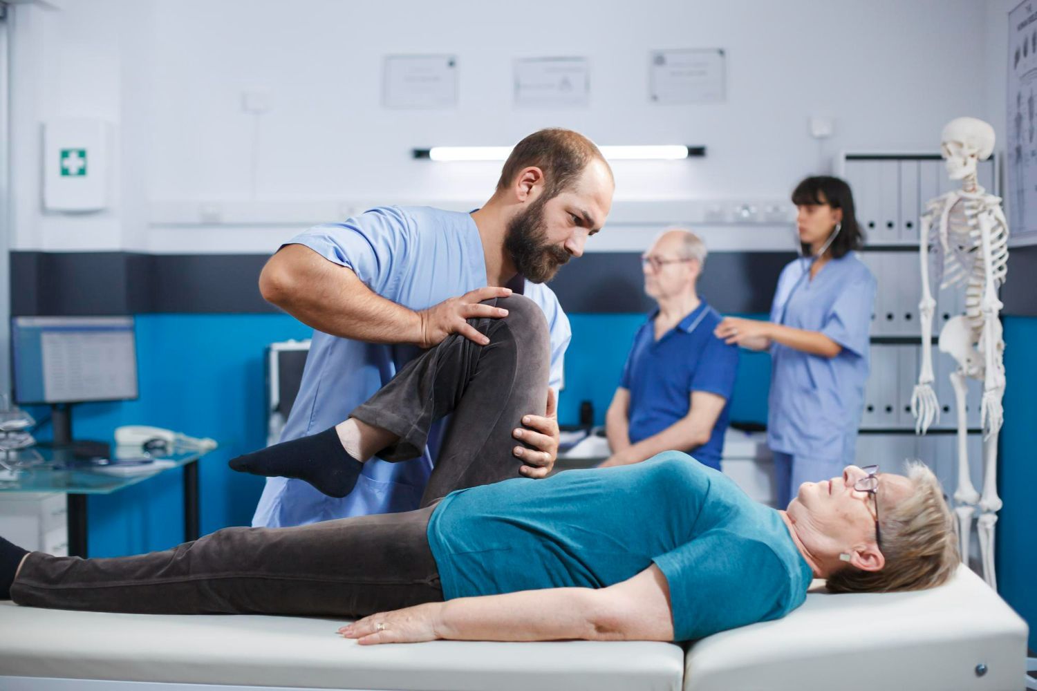 Physiotherapist examining patient's leg in clinic; another patient and nurse in background.