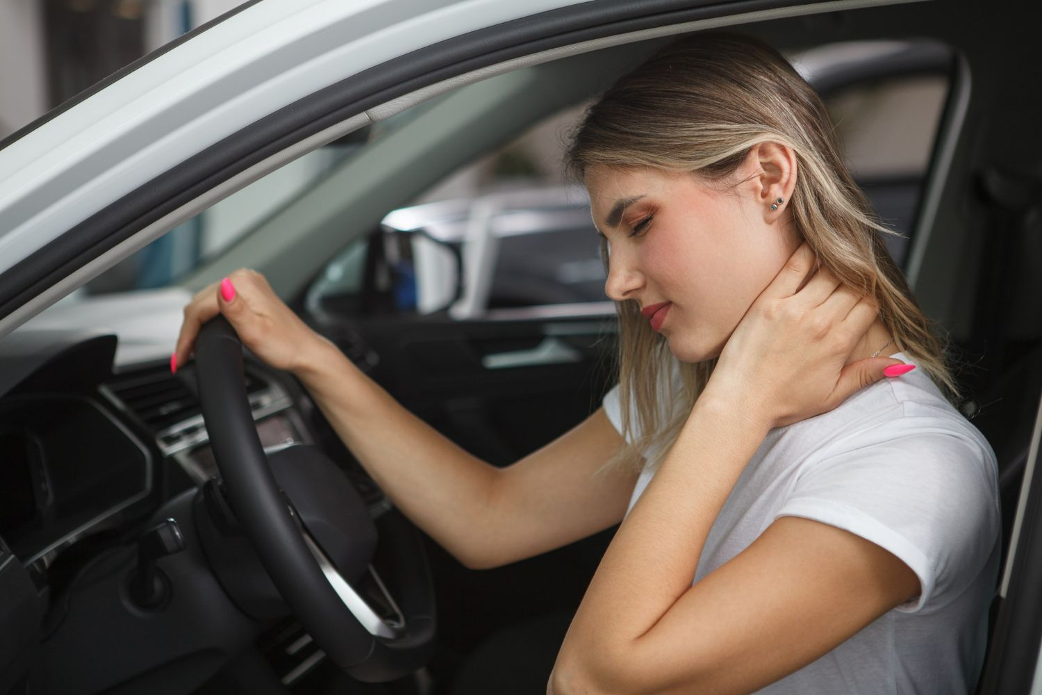A person sitting in a car seat holding their neck and looking distressed.