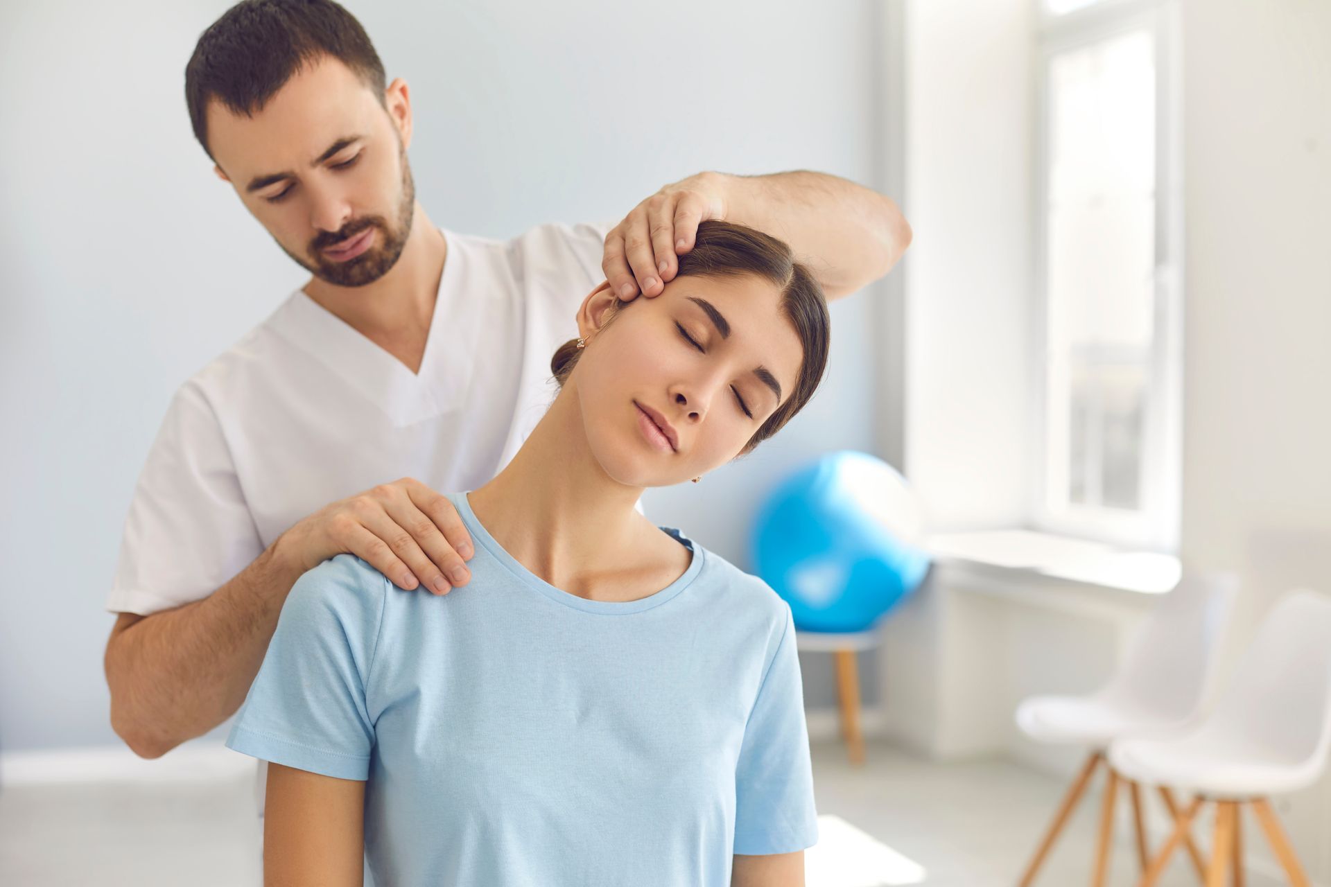 Man adjusting a person's neck. Person in blue shirt has eyes closed, standing in a room with a blue exercise ball.