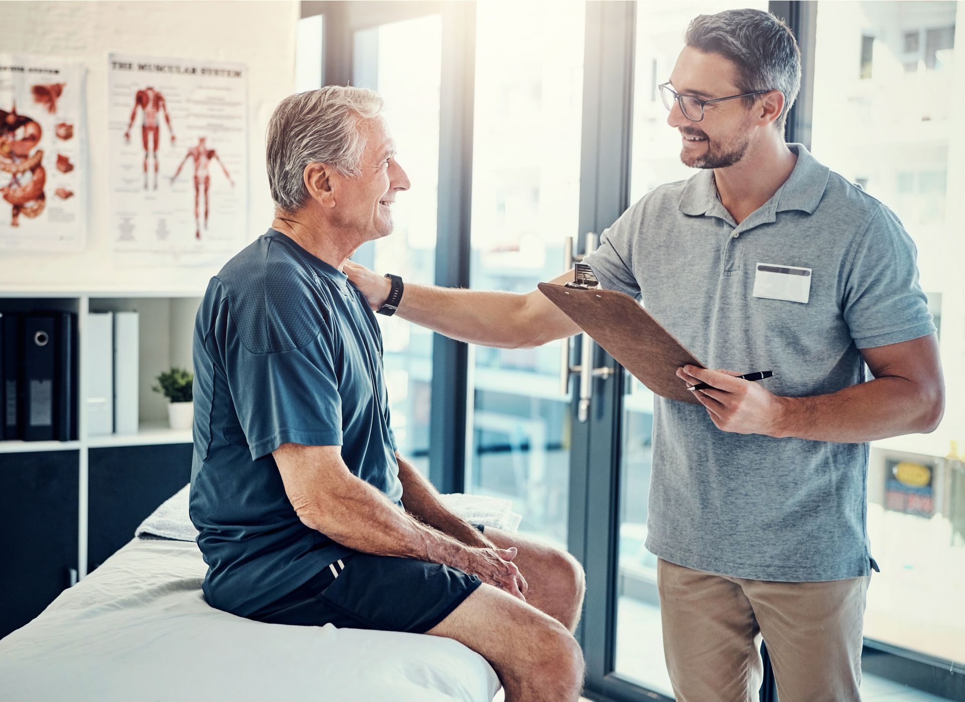 A man is sitting on a bed talking to a doctor who is holding a clipboard.