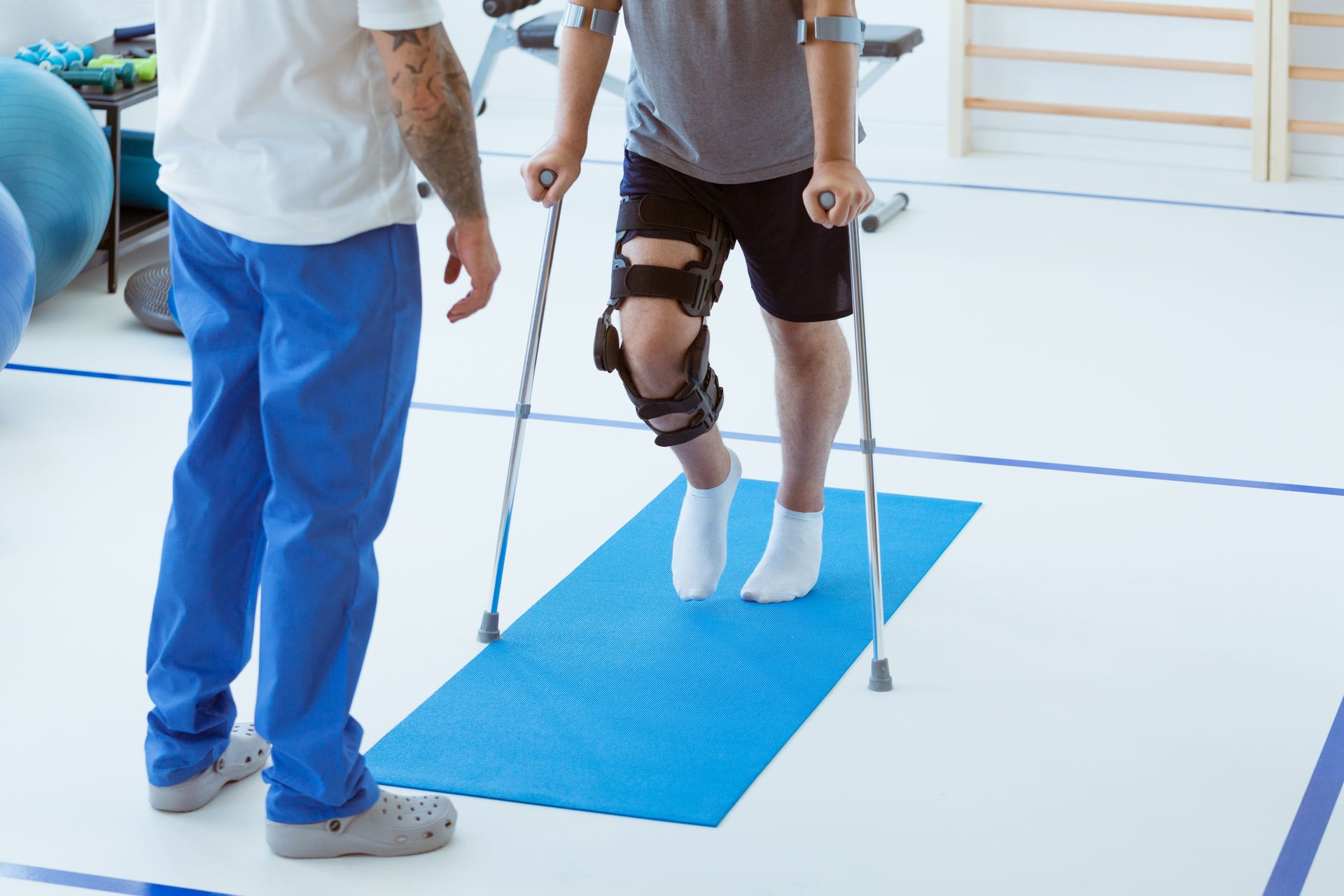 A man with a knee brace is using crutches to walk on a blue mat.