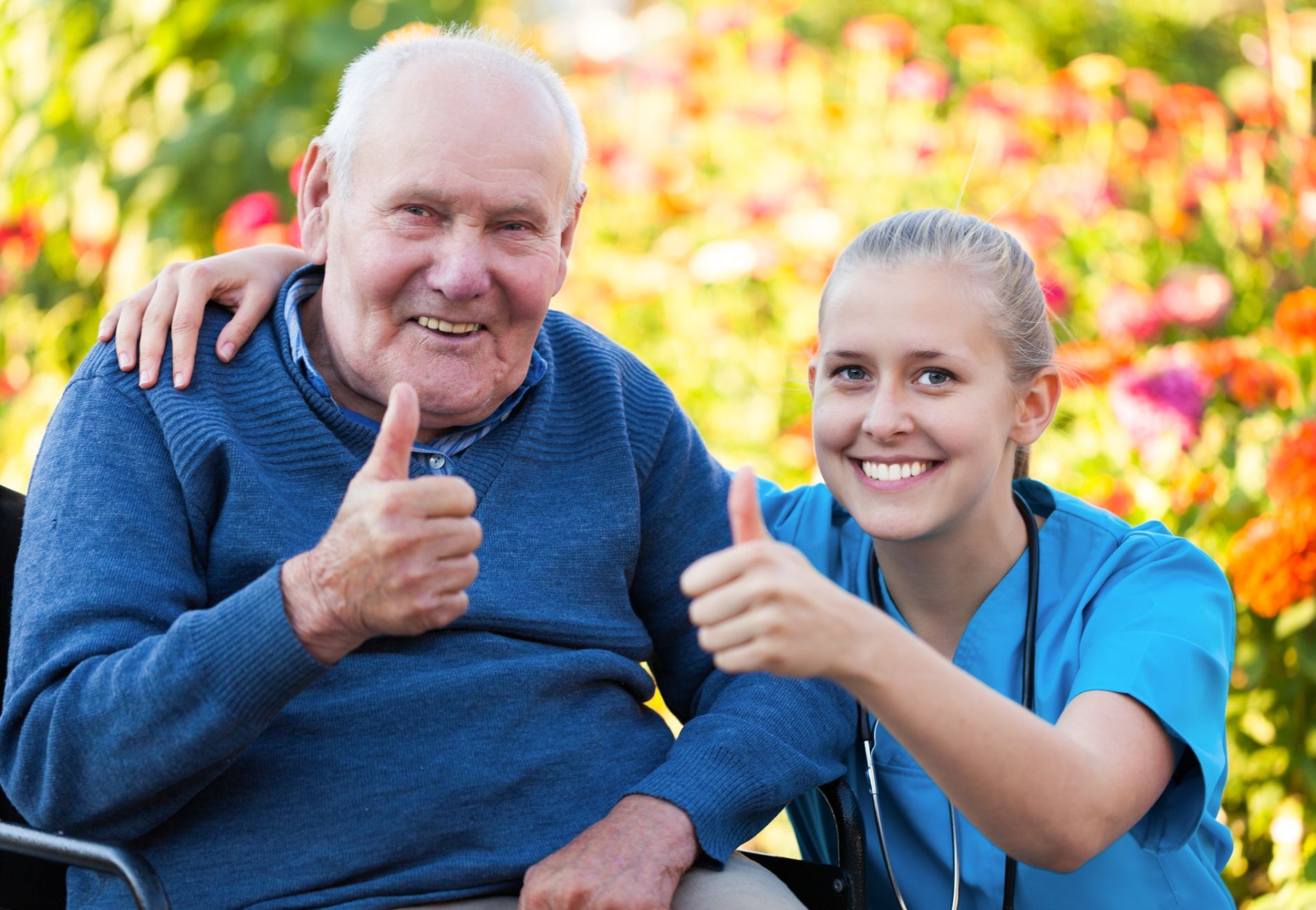 Elderly man and female caregiver smiling and giving thumbs up outdoors.