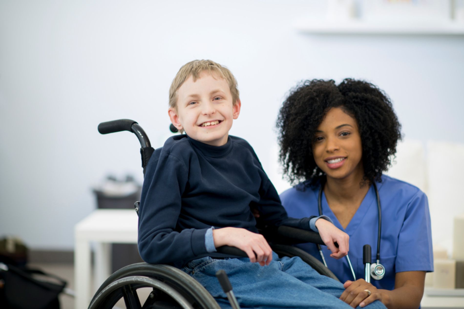 Boy in wheelchair smiles with a nurse in blue scrubs, indoors.