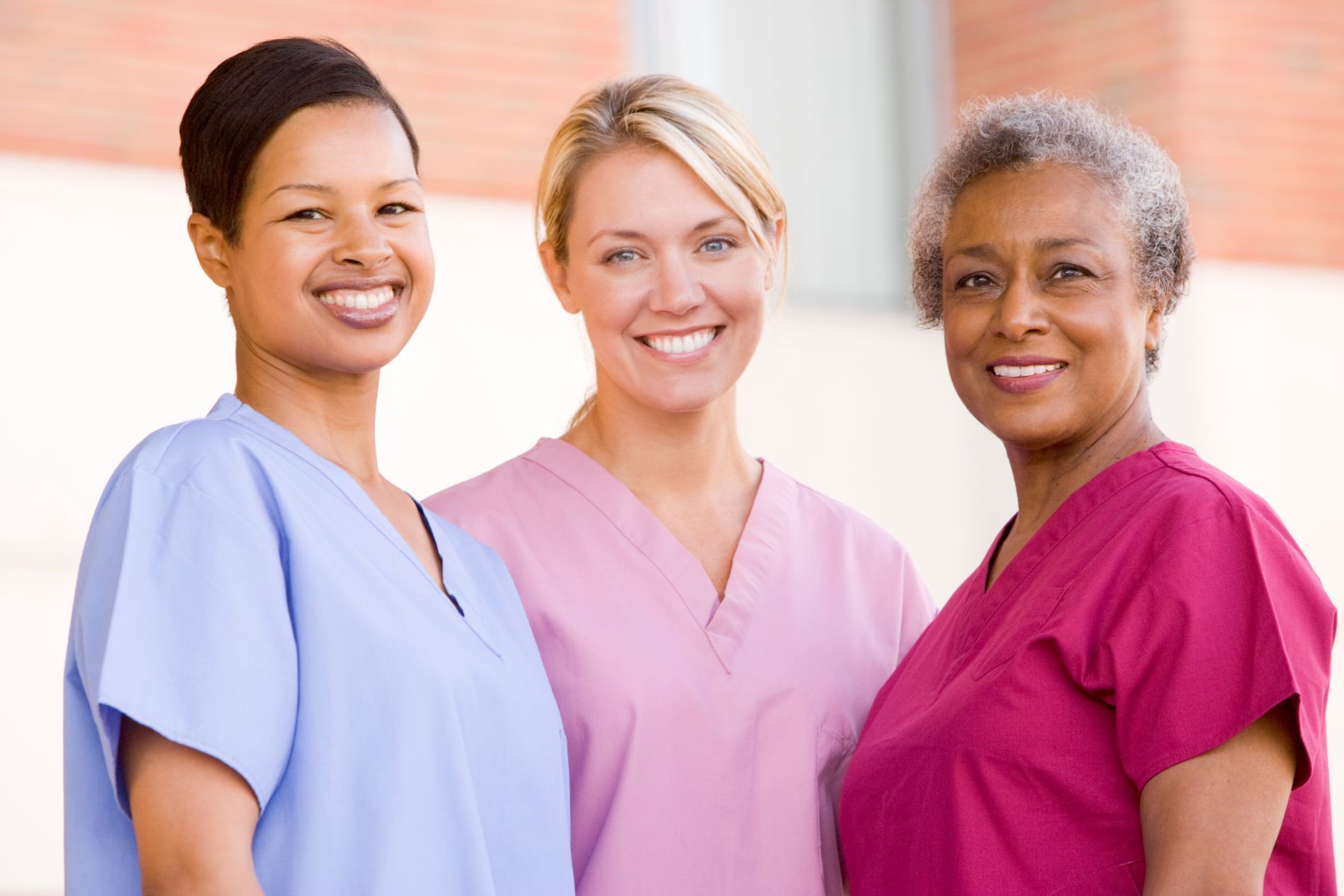 Three smiling nurses in scrubs of blue, pink, and red, standing outside a building.
