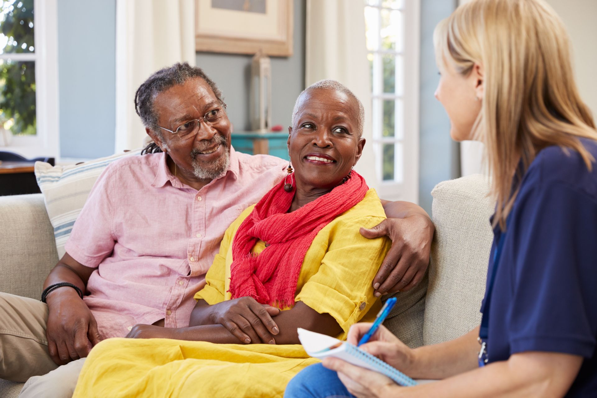 An elderly couple on a couch consult with a caregiver in a living room setting.