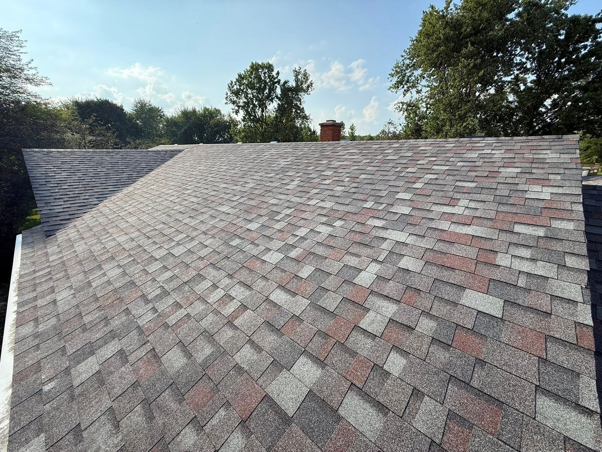 View of a multi-colored shingled roof with a chimney, against a blue sky, trees in the background.