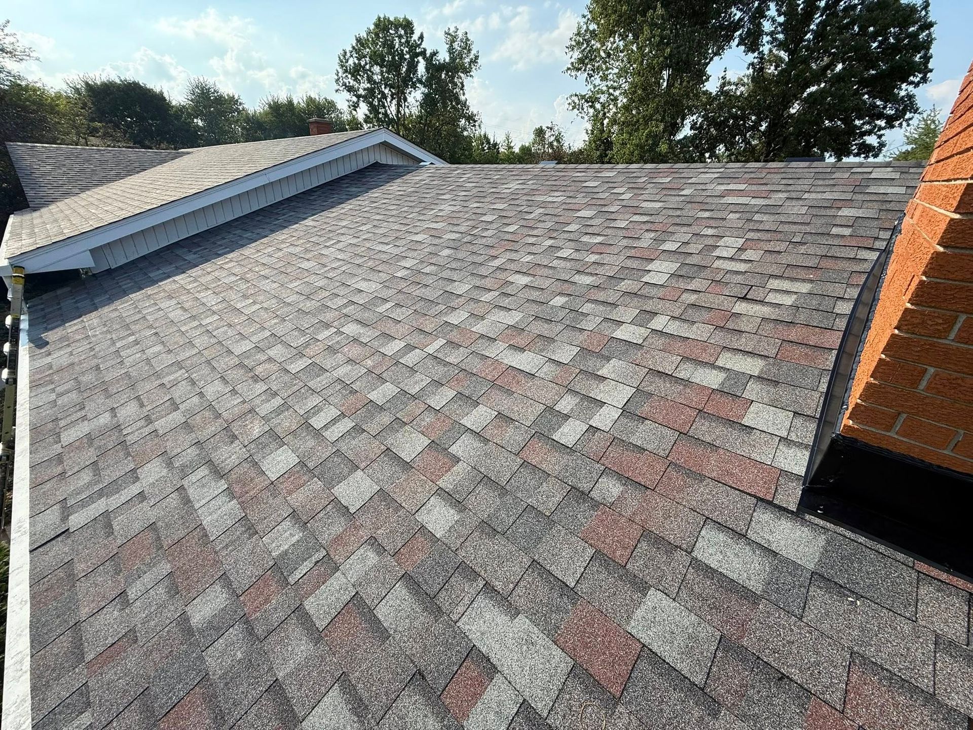 Asphalt shingle roof with gray, red, and white patches, chimney on the right, trees in the background.