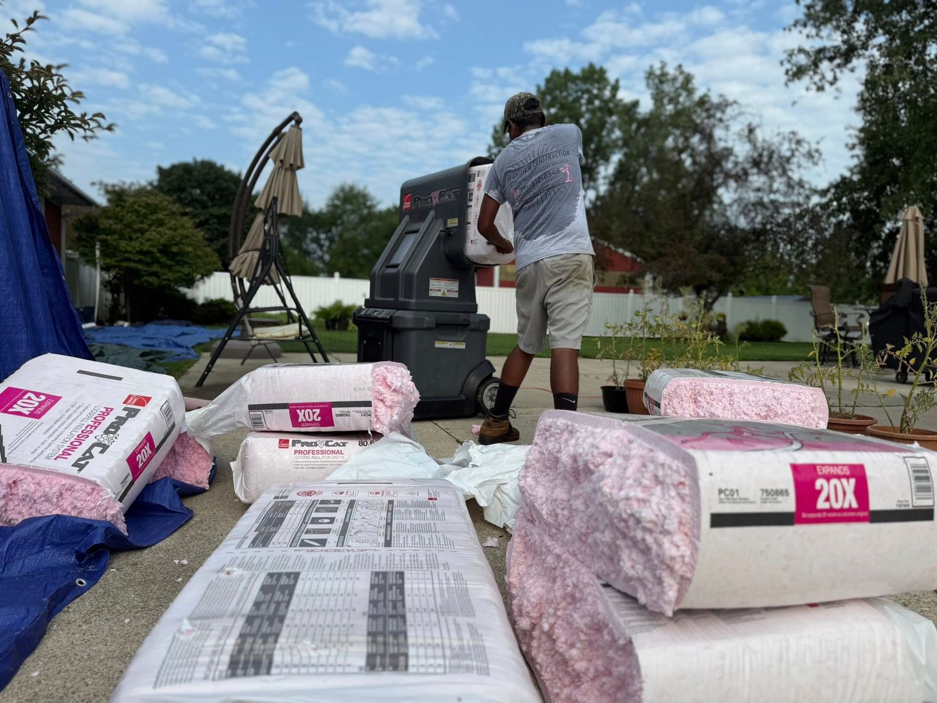 Person loading insulation into a machine outdoors. Insulation bales and backyard visible.