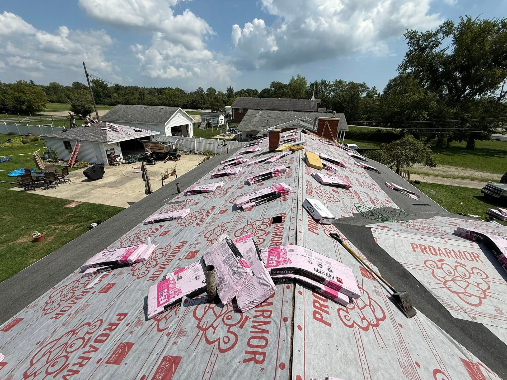Rooftop under construction with pink protective underlayment and partially installed shingles. Blue sky, houses in background.