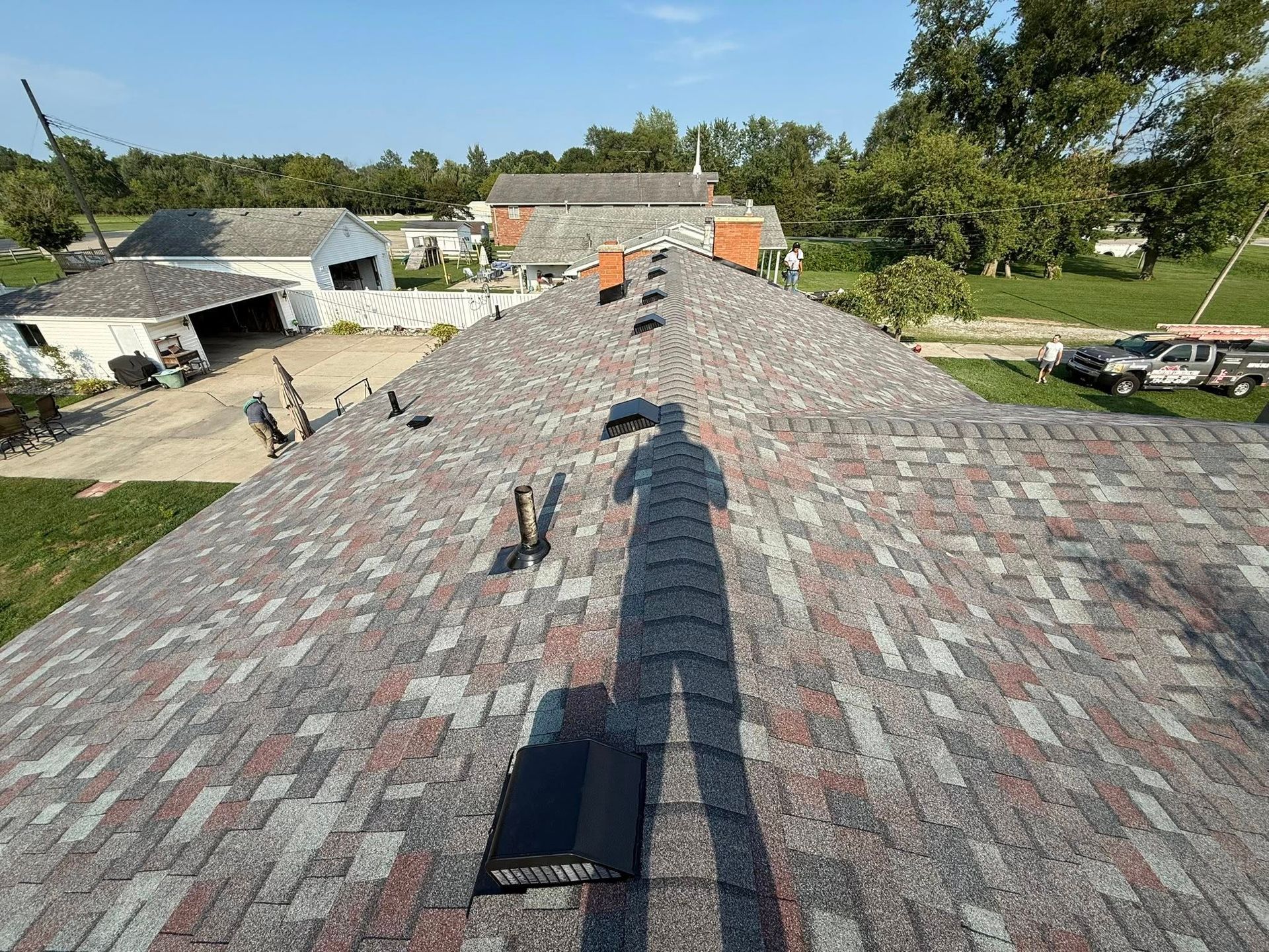 Shadow of a person on a roof with gray, red, and brown shingles, overlooking buildings and trees on a sunny day.
