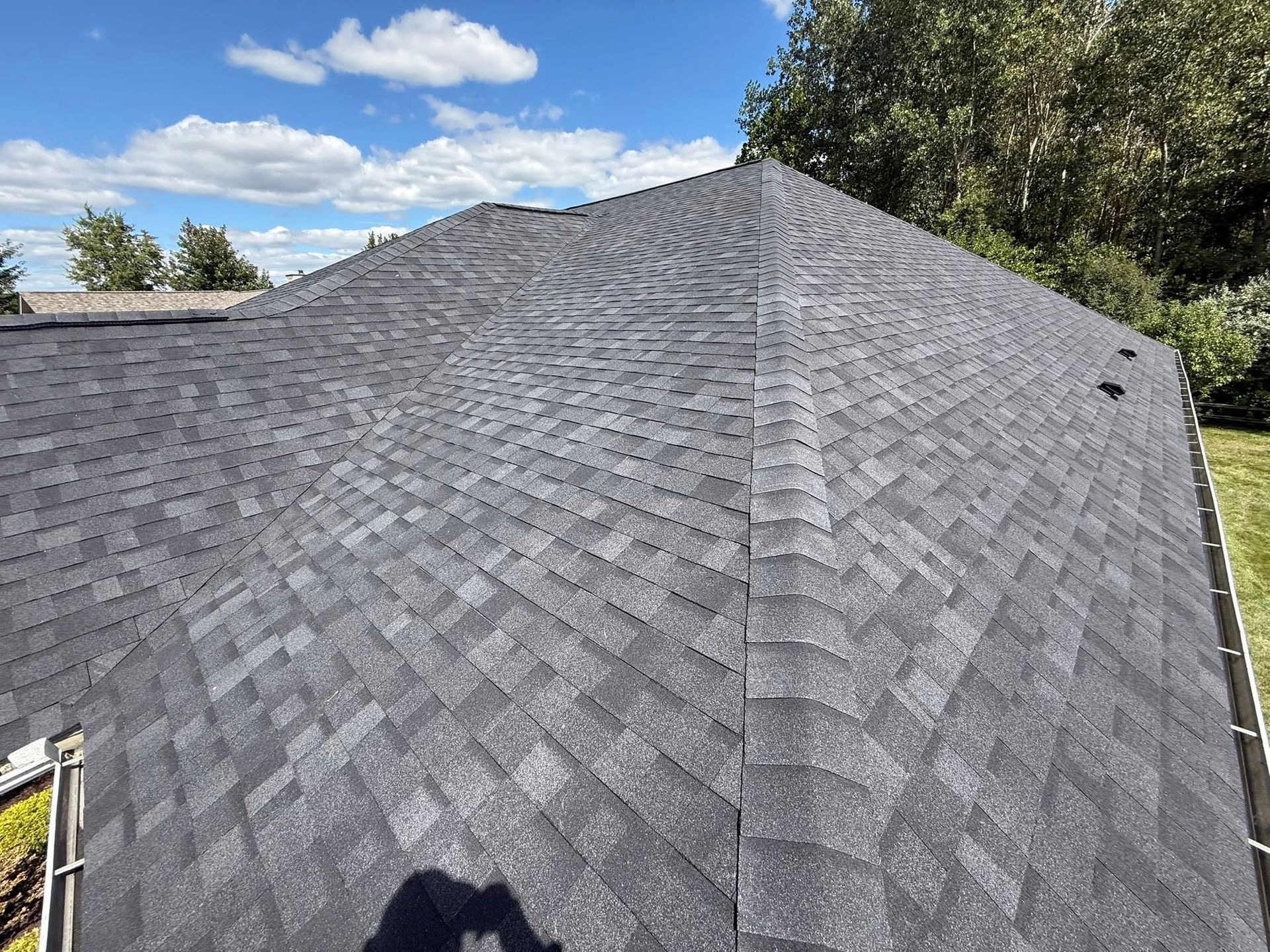 Dark gray asphalt shingle roof on a house, under a partly cloudy blue sky.
