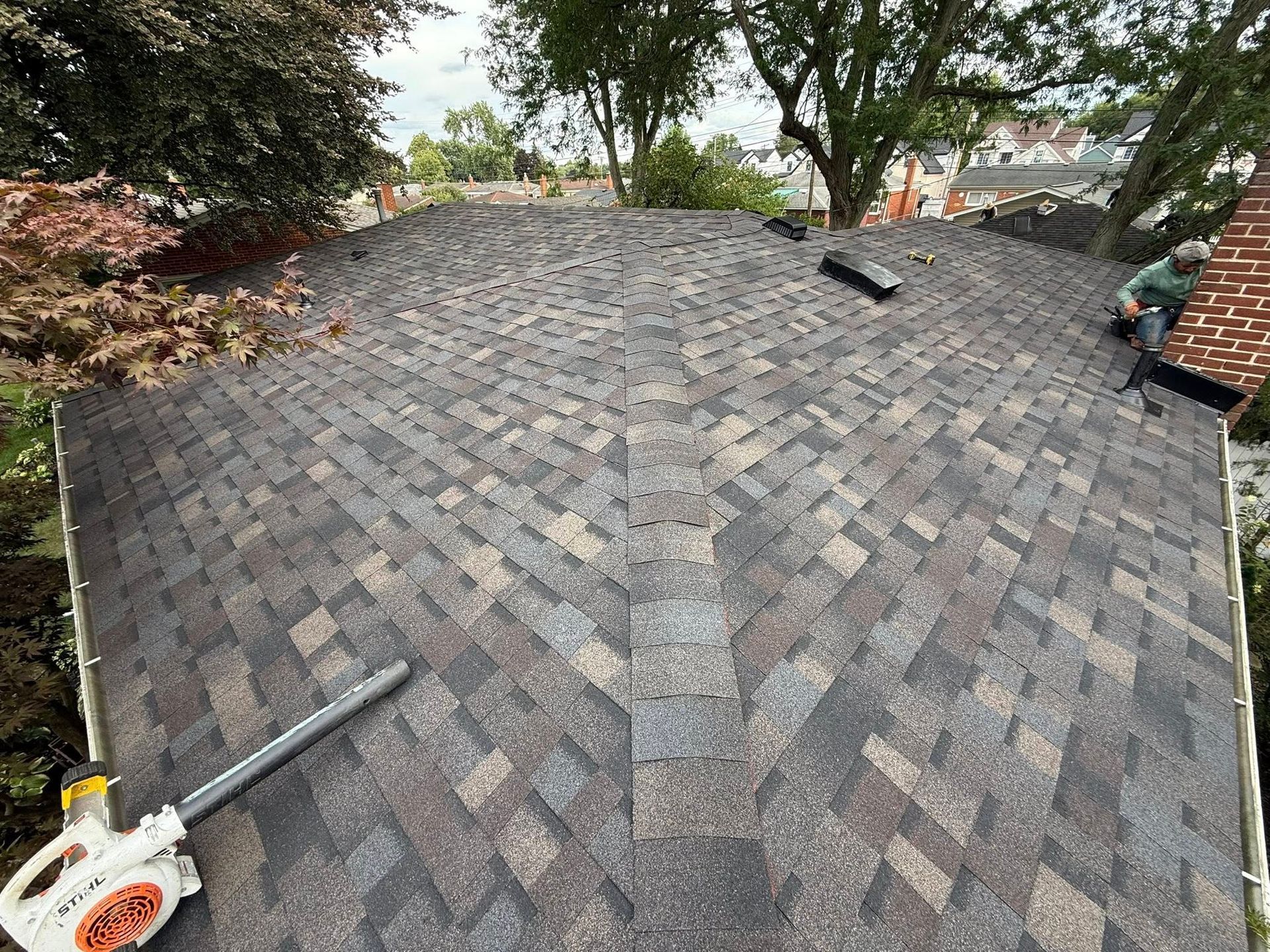 Newly shingled roof with brown and grey shingles; worker near chimney. Leaf blower in the foreground.