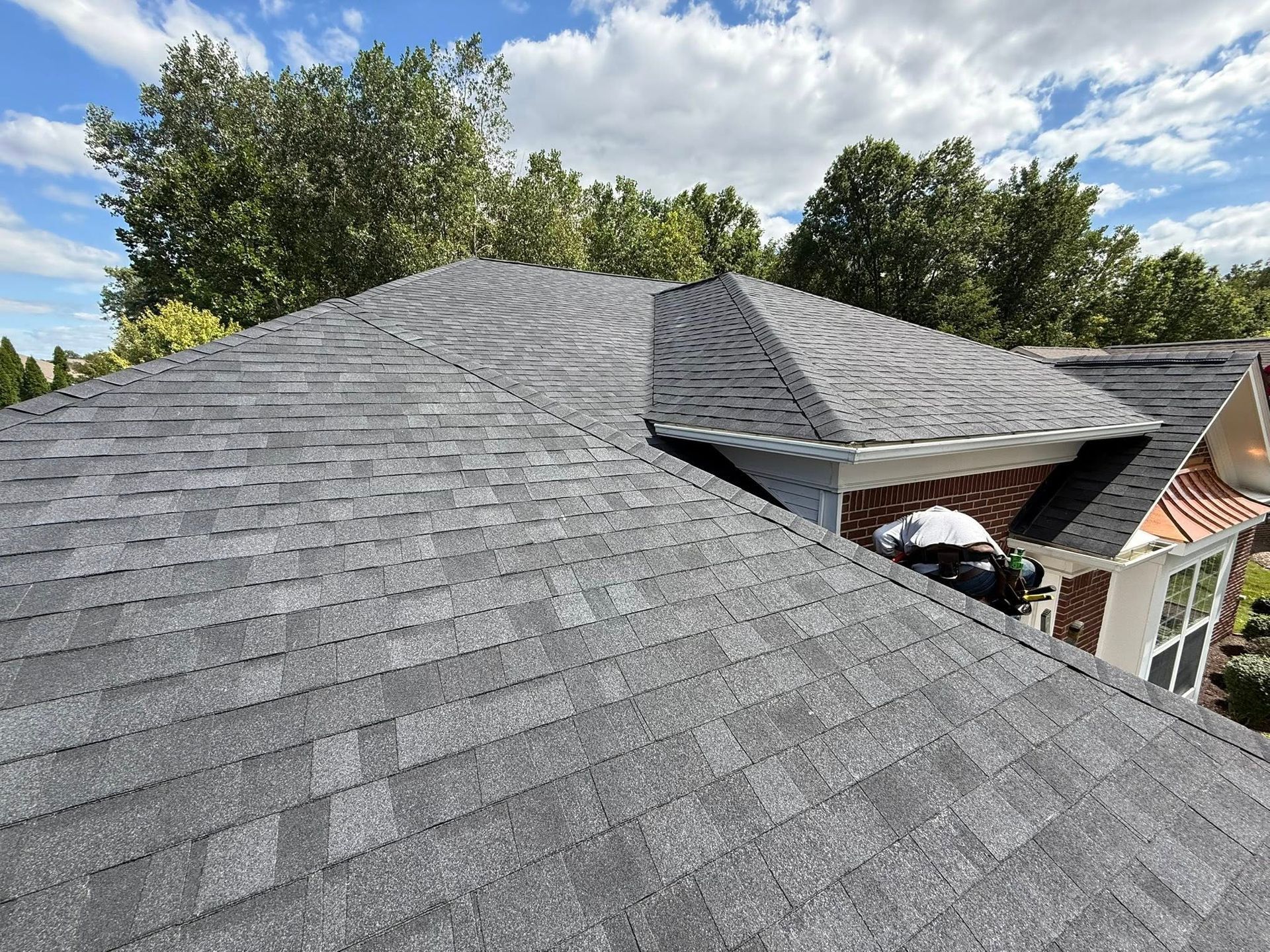 Gray asphalt shingle roof on a house, with a small section of a dormer, against a cloudy blue sky.