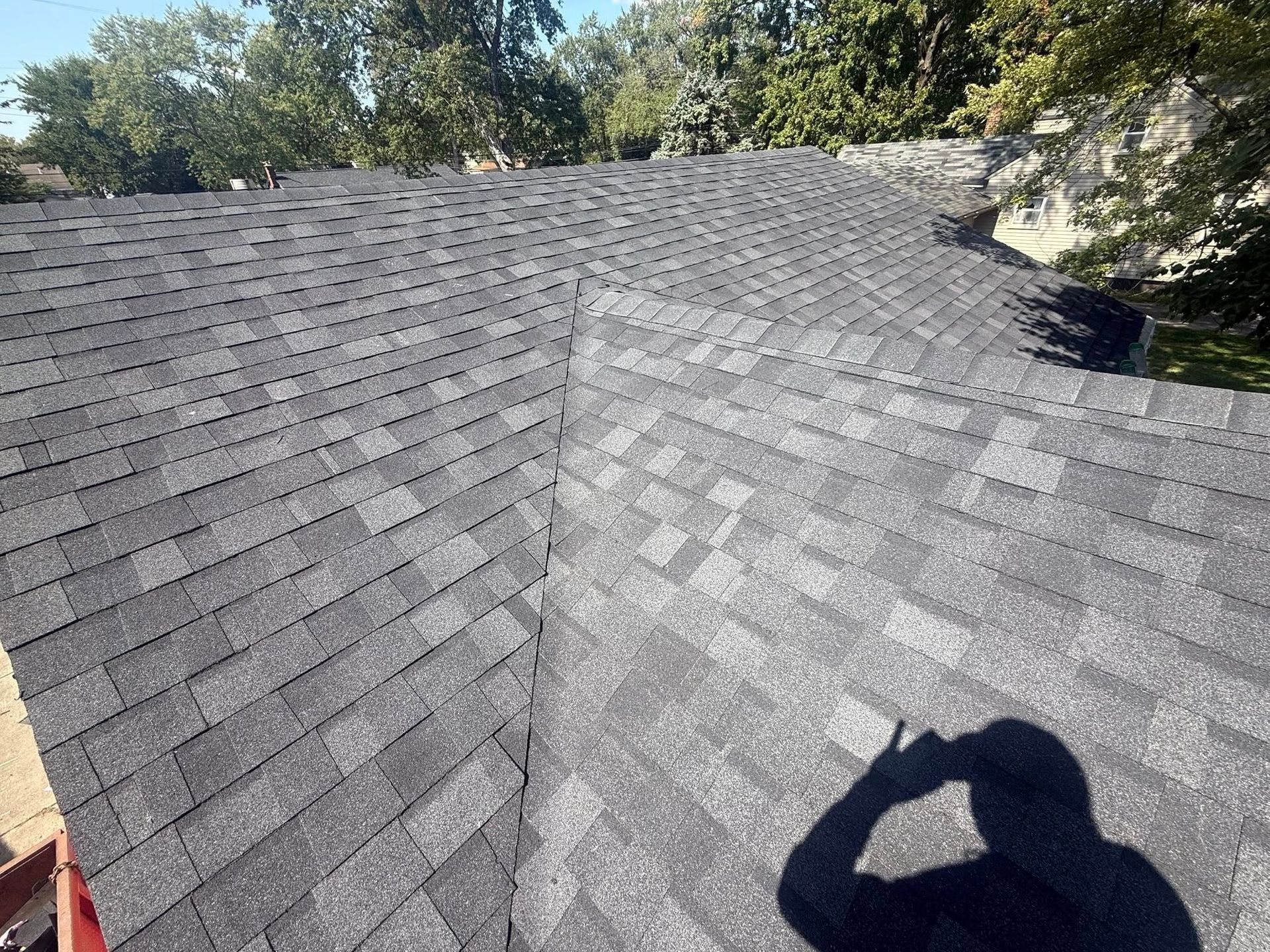 Dark gray shingled roof with trees in background, sunny day. Shadow of person taking photo.