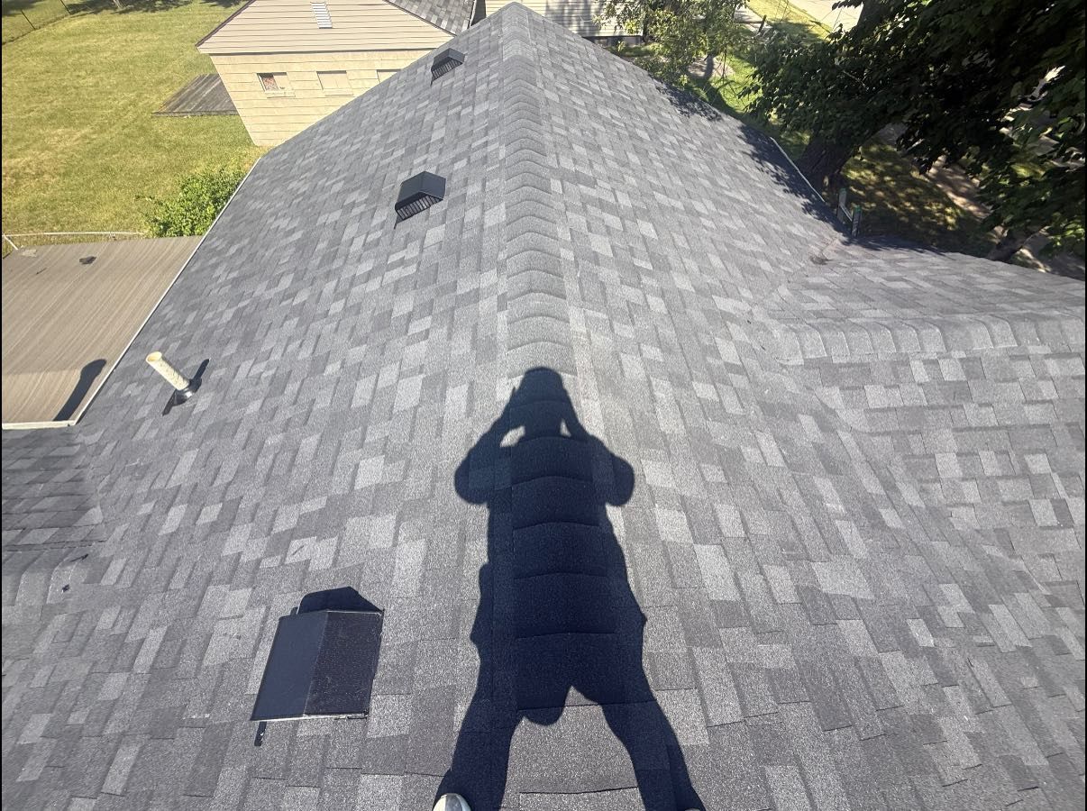 Person's shadow on a gray asphalt shingle roof. Three vents visible. Green vegetation and building in background.