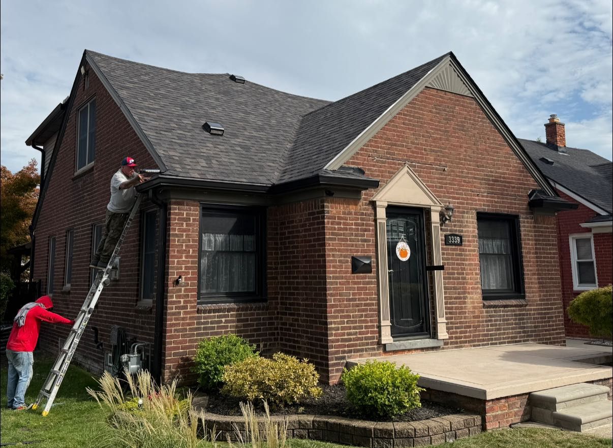 Two people on ladder cleaning gutters of a brick house with black trim and a dark roof.