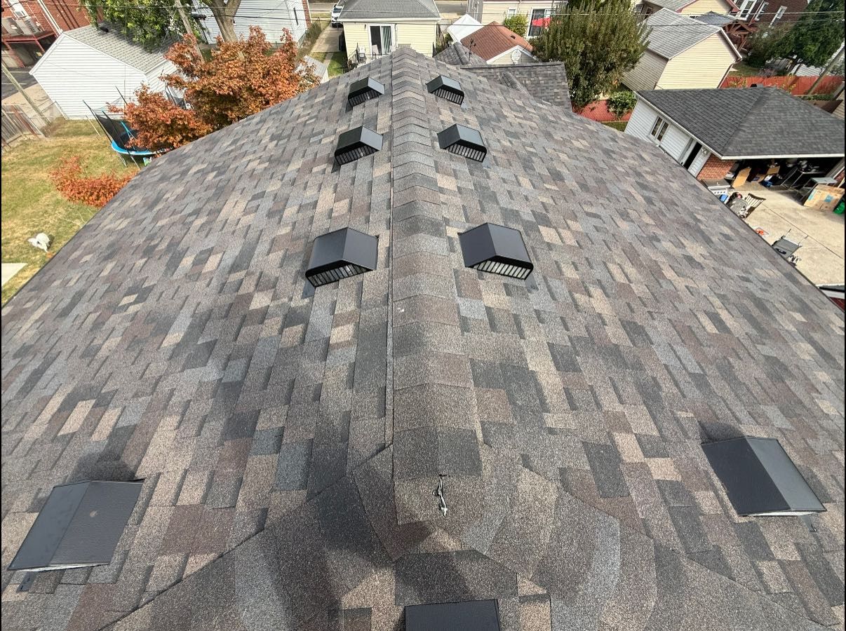 Overhead view of a roof with asphalt shingles and black vents. Houses and trees are visible in the background.