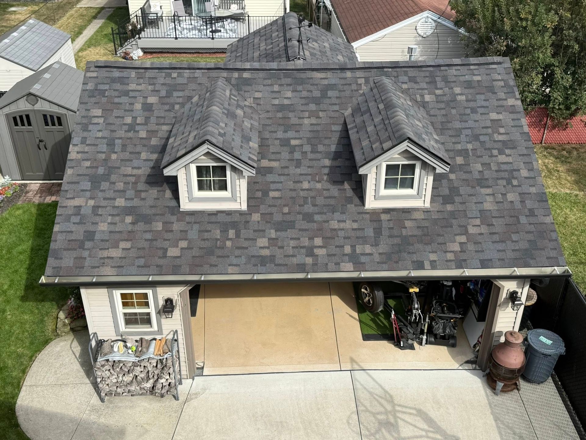 Garage with dark gray shingle roof, two dormer windows, tan garage door. Lawn and shed visible.