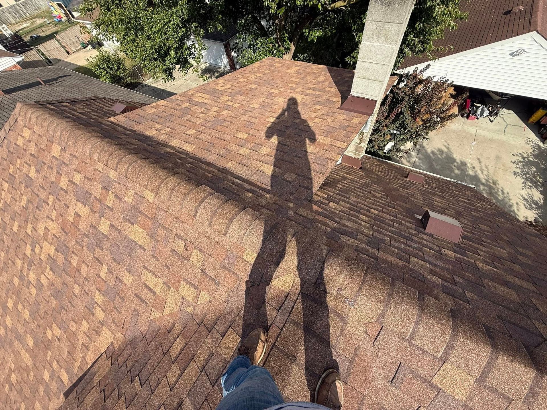 Person's shadow on a brown shingled roof, trees and other rooftops visible in background.