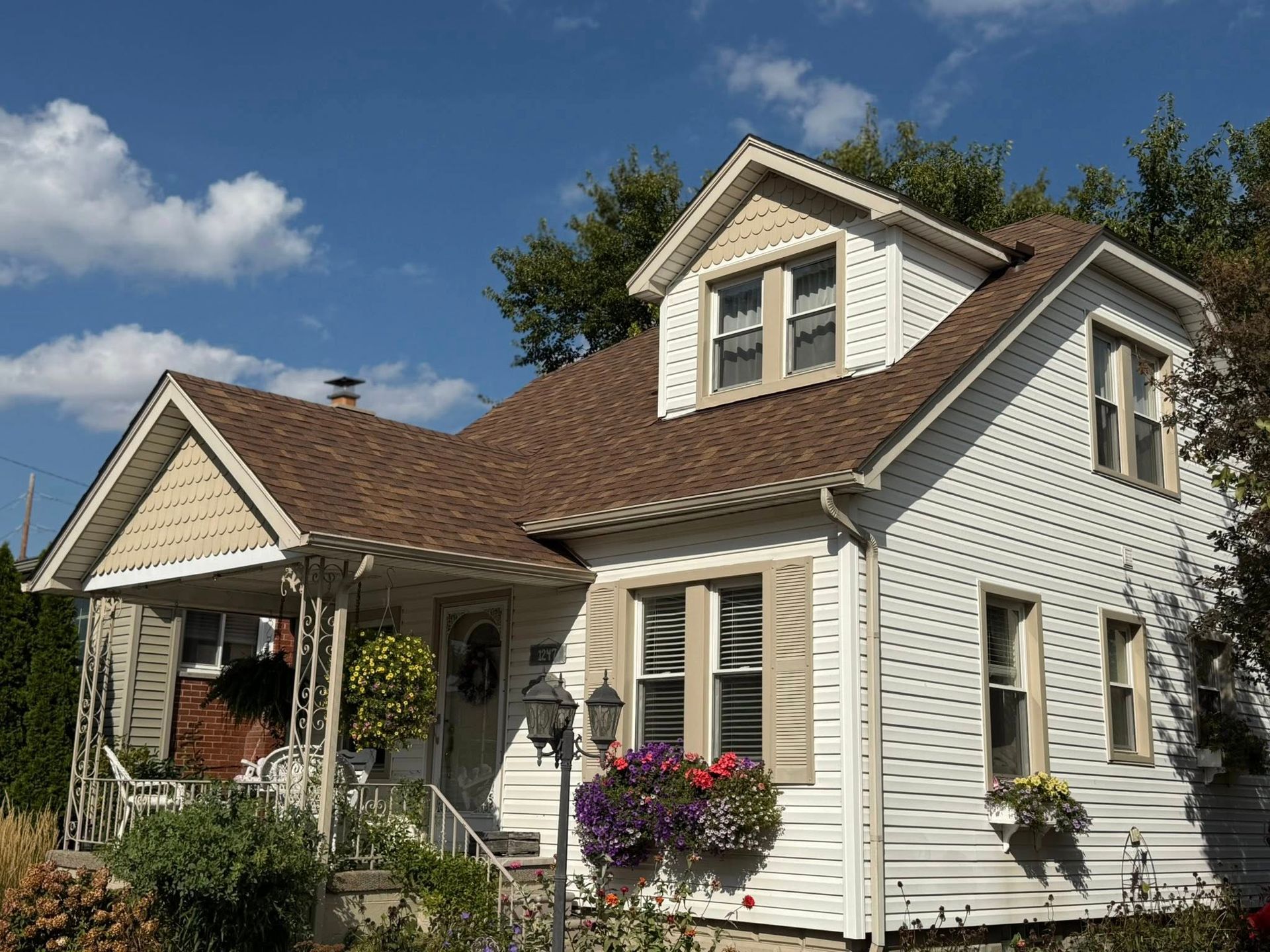White house with brown roof and beige trim, under a blue sky. Flowers in window boxes.