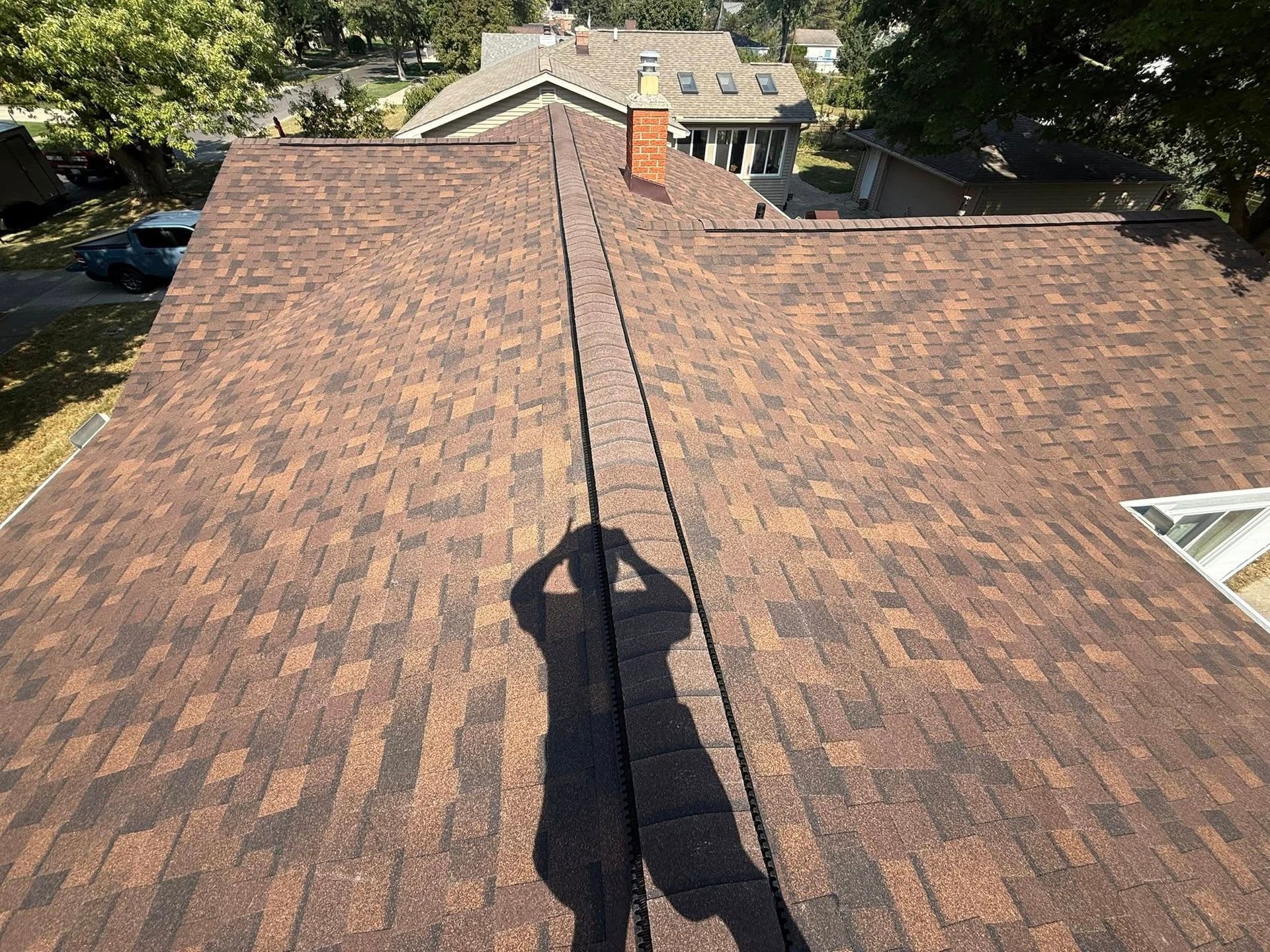 Brown asphalt shingle roof on a house, with a long shadow of a person taking a photo.