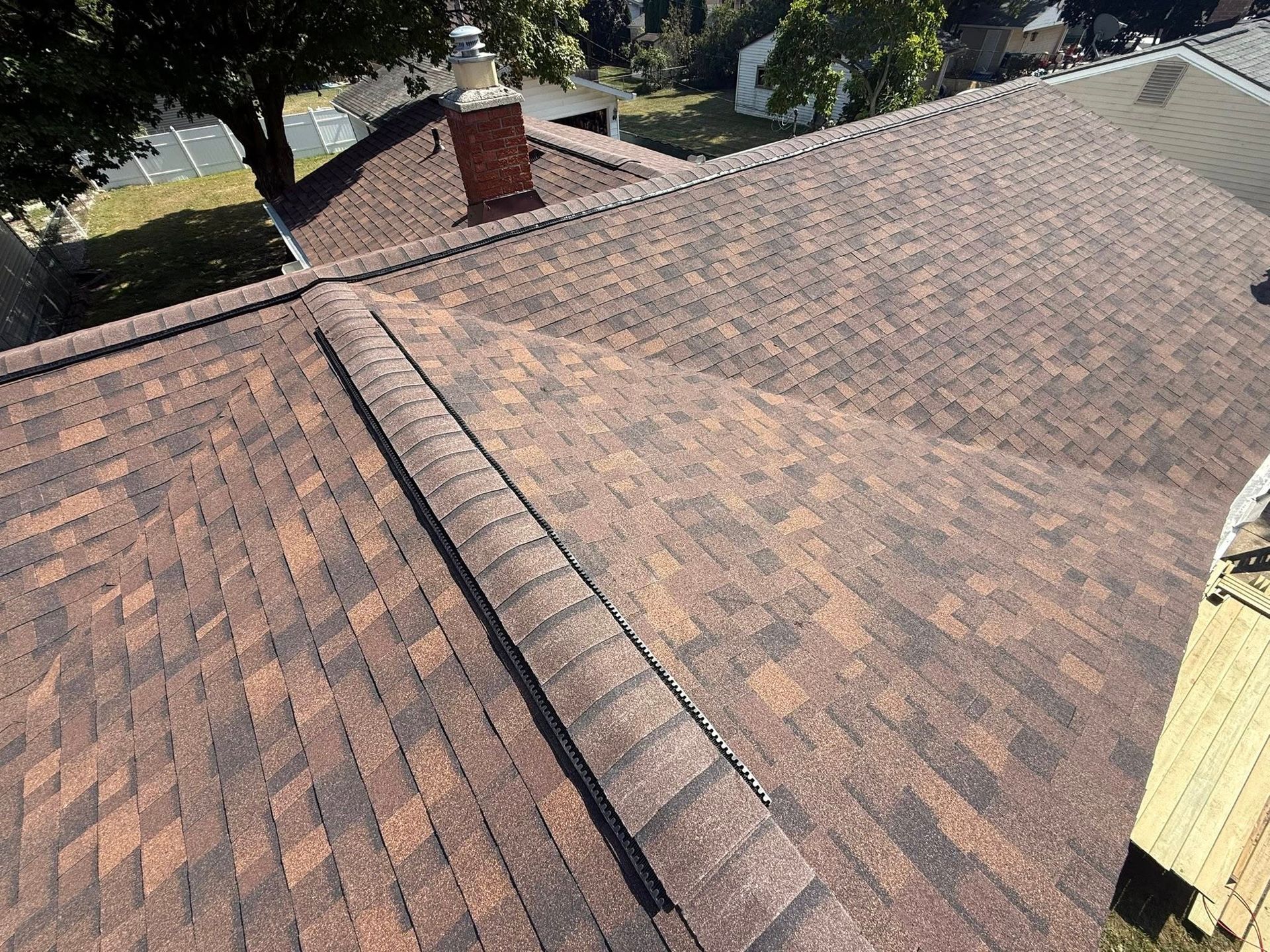 Brown asphalt shingle roof, with a chimney and ridge cap visible. Overlooking a yard and trees.