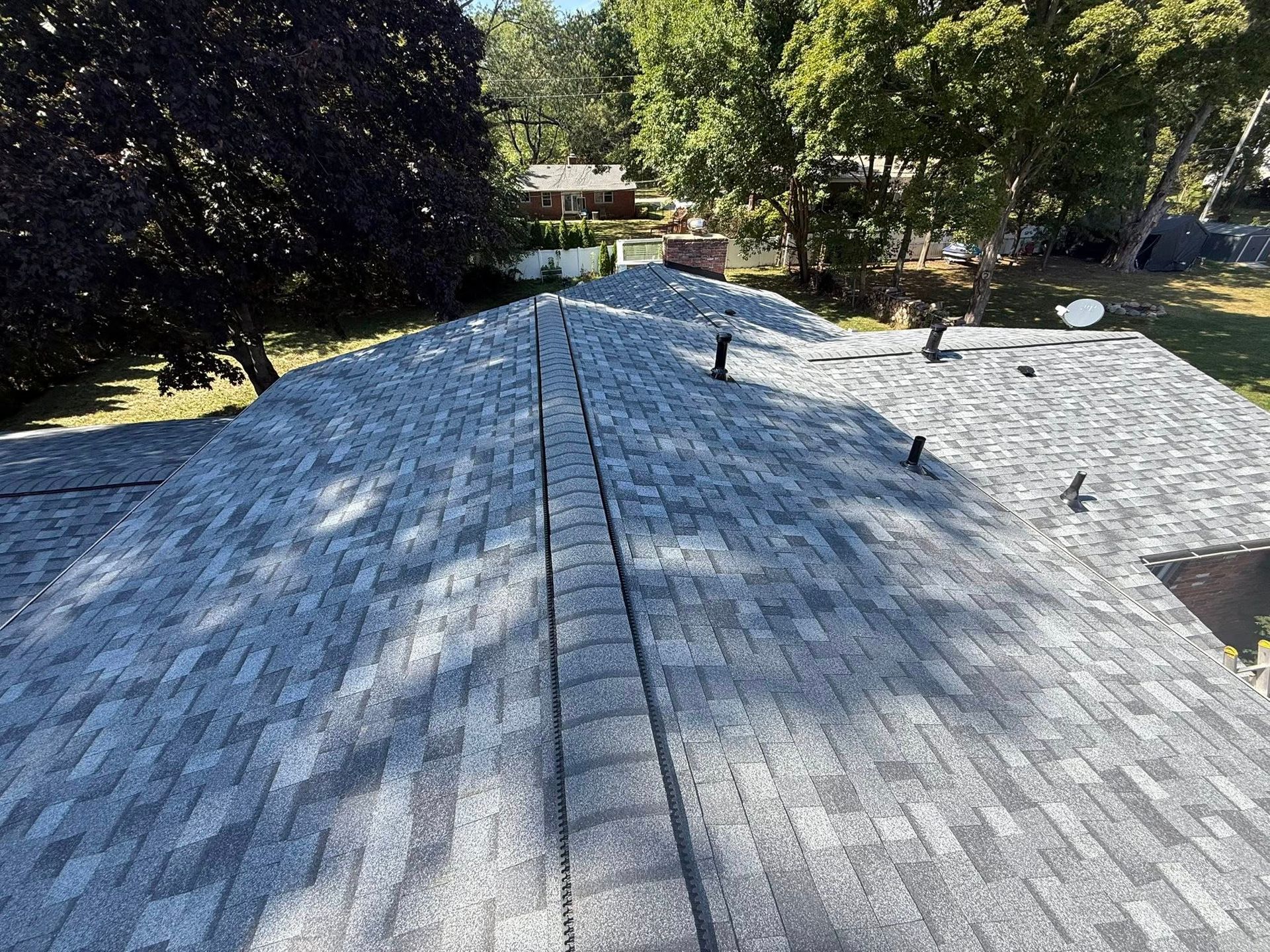Gray asphalt shingle roof with vents against a backdrop of trees and a sunny sky.