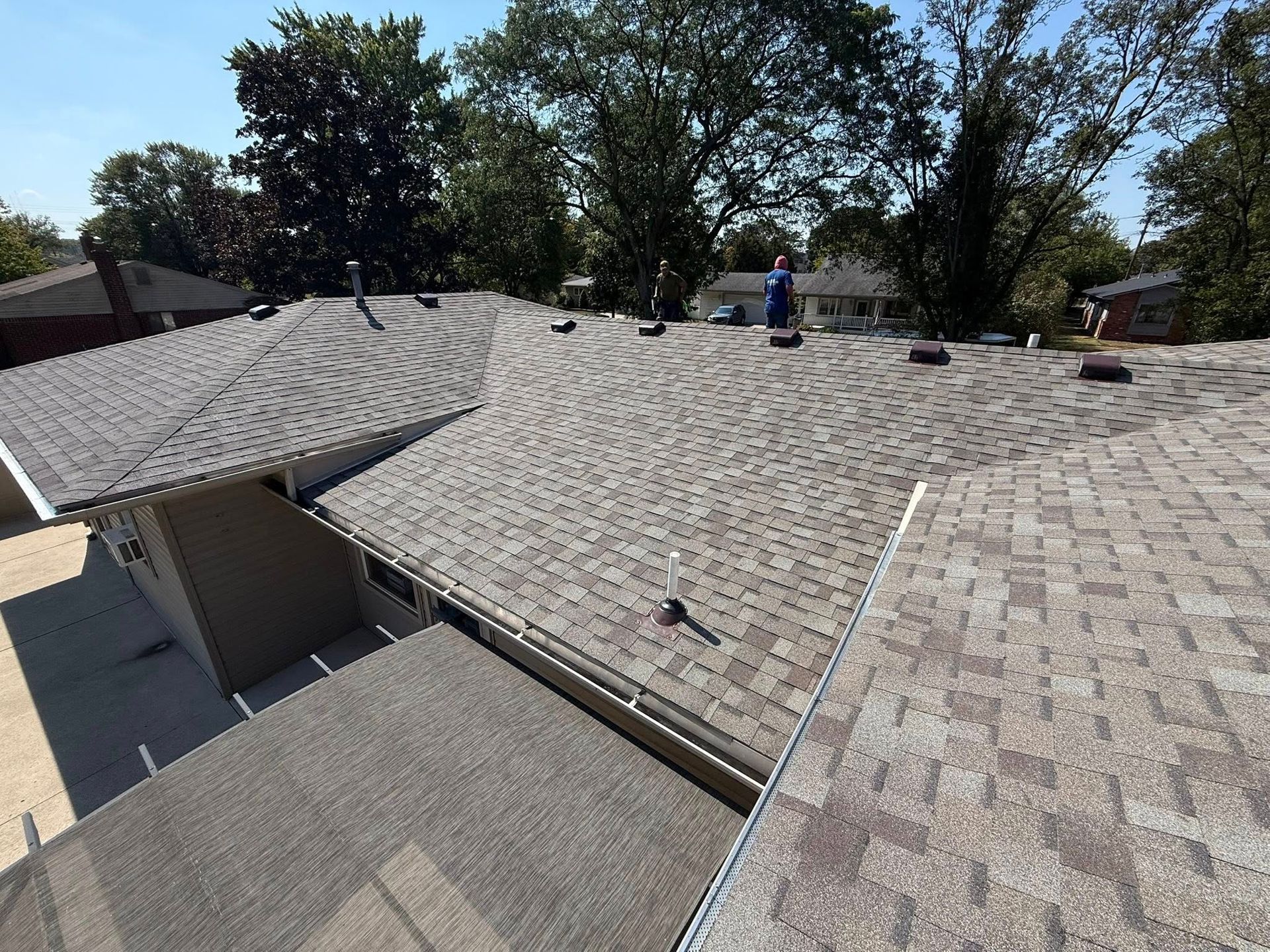 View of a gray shingled roof with a person standing on it, trees in the background, sunny day.