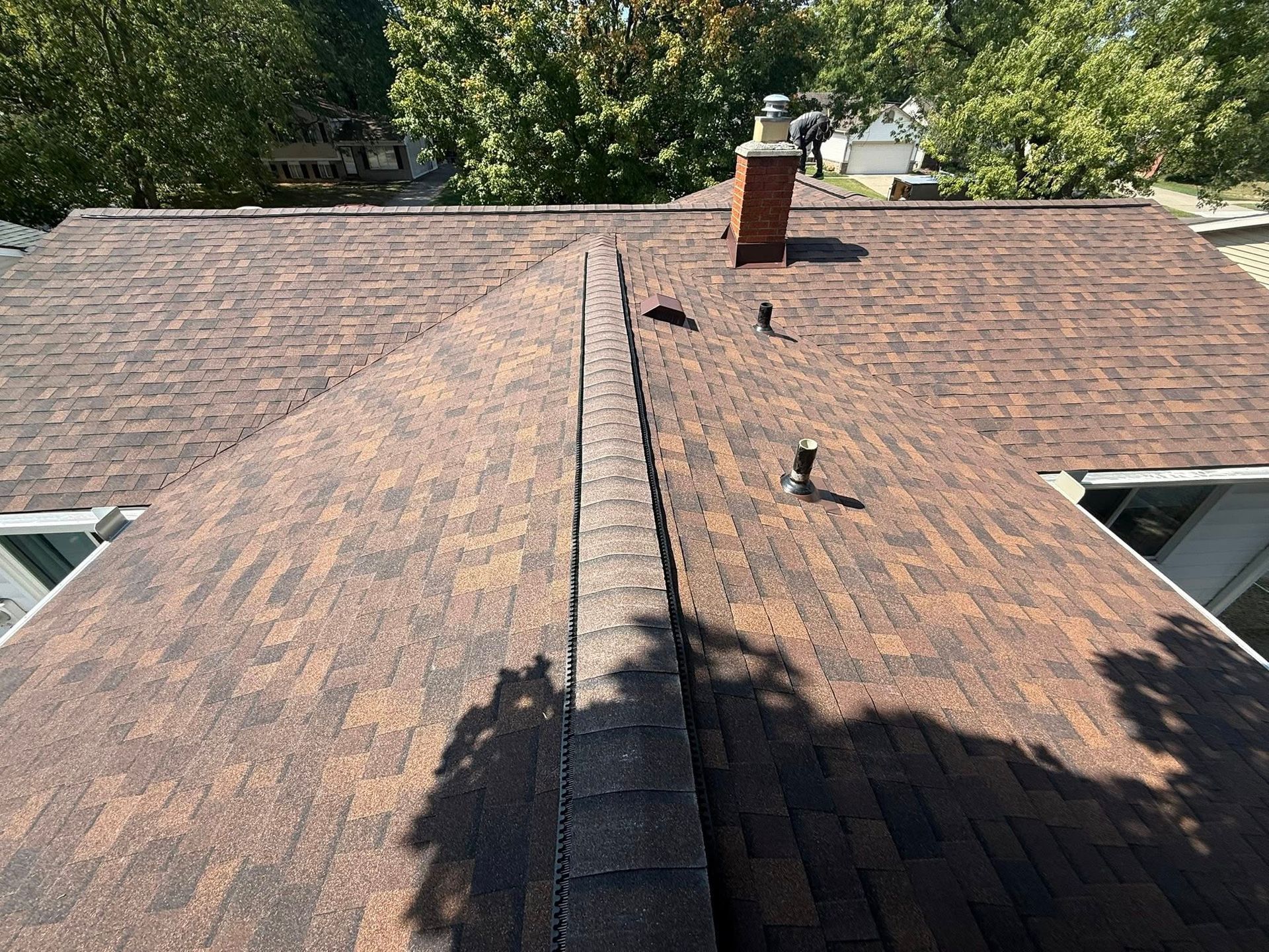 Brown shingle roof with a brick chimney and vent pipe, viewed from above on a sunny day.