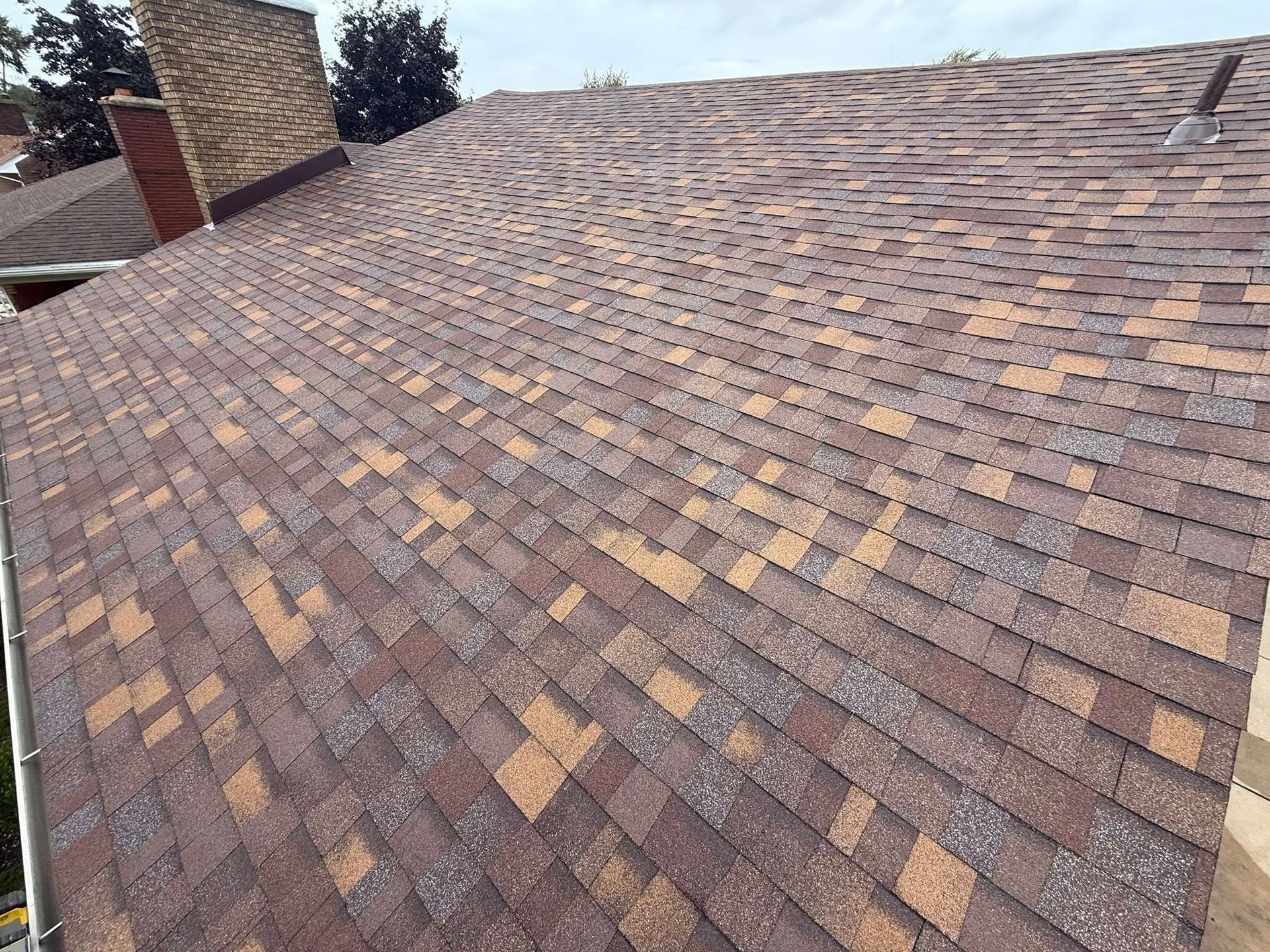 Brown shingle roof with varying shades, angled view, chimney and gutter visible, overcast sky.