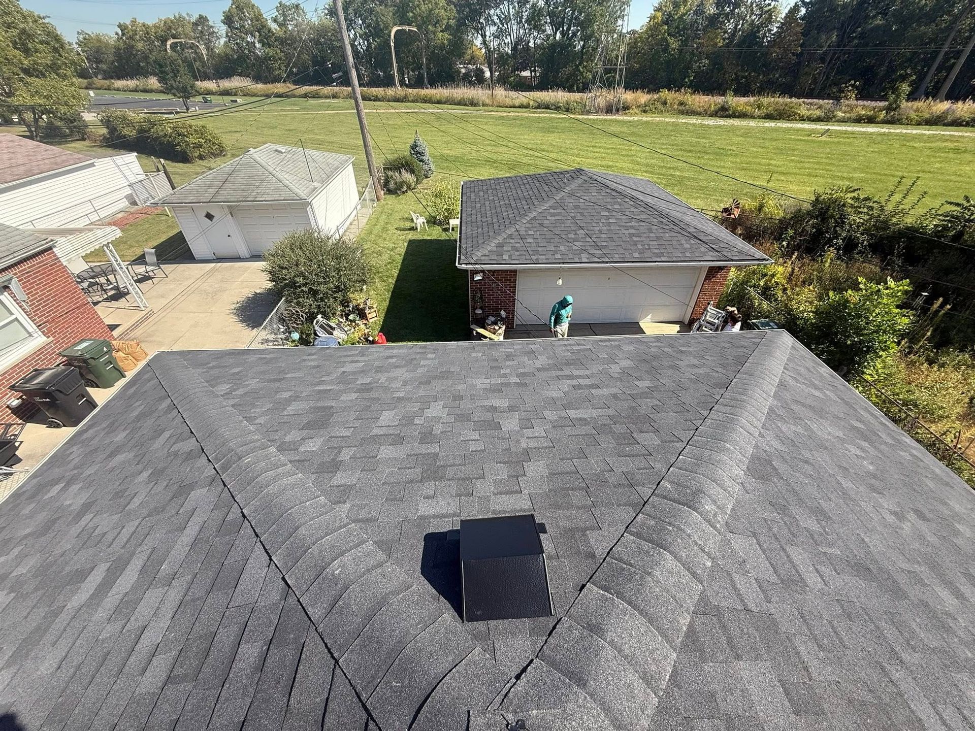 View of a dark gray shingled roof with a chimney, houses, and green field in the background.