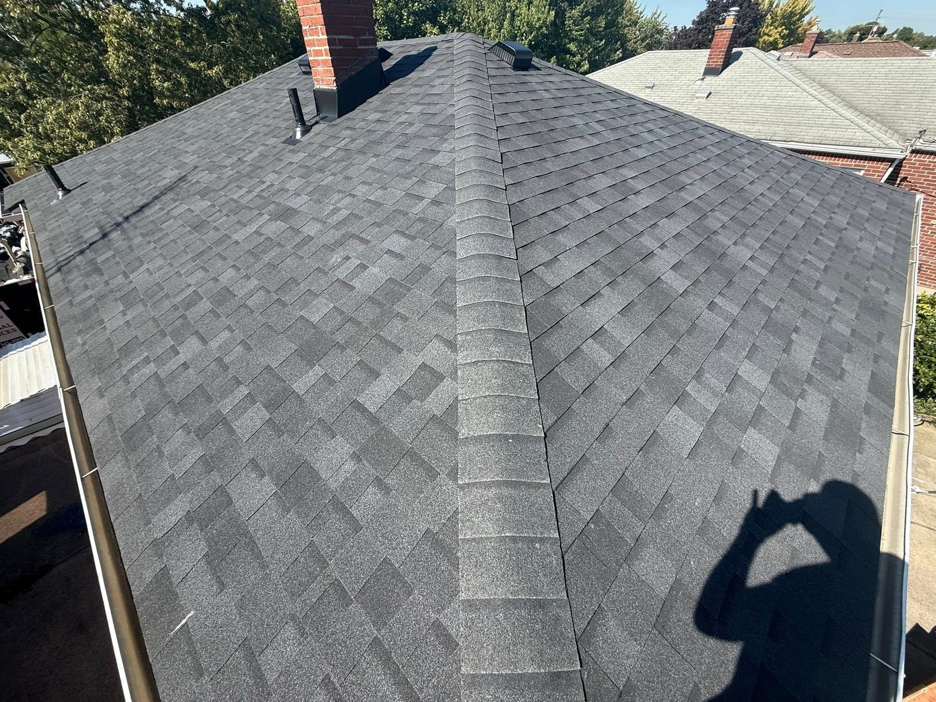 Dark gray asphalt shingle roof on a house with a brick chimney, seen from above on a sunny day.
