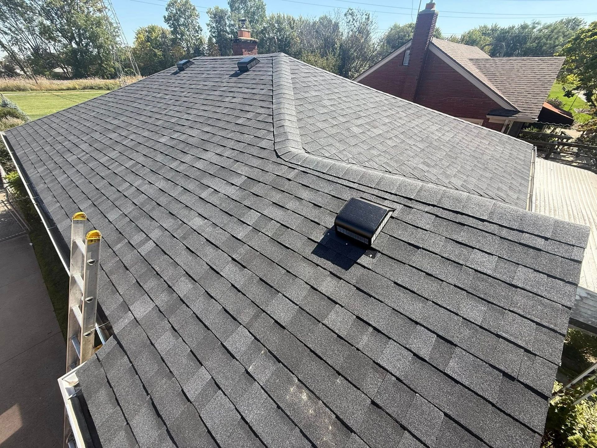 A rooftop with dark gray shingles under a clear, sunny sky. A ladder rests on the side.