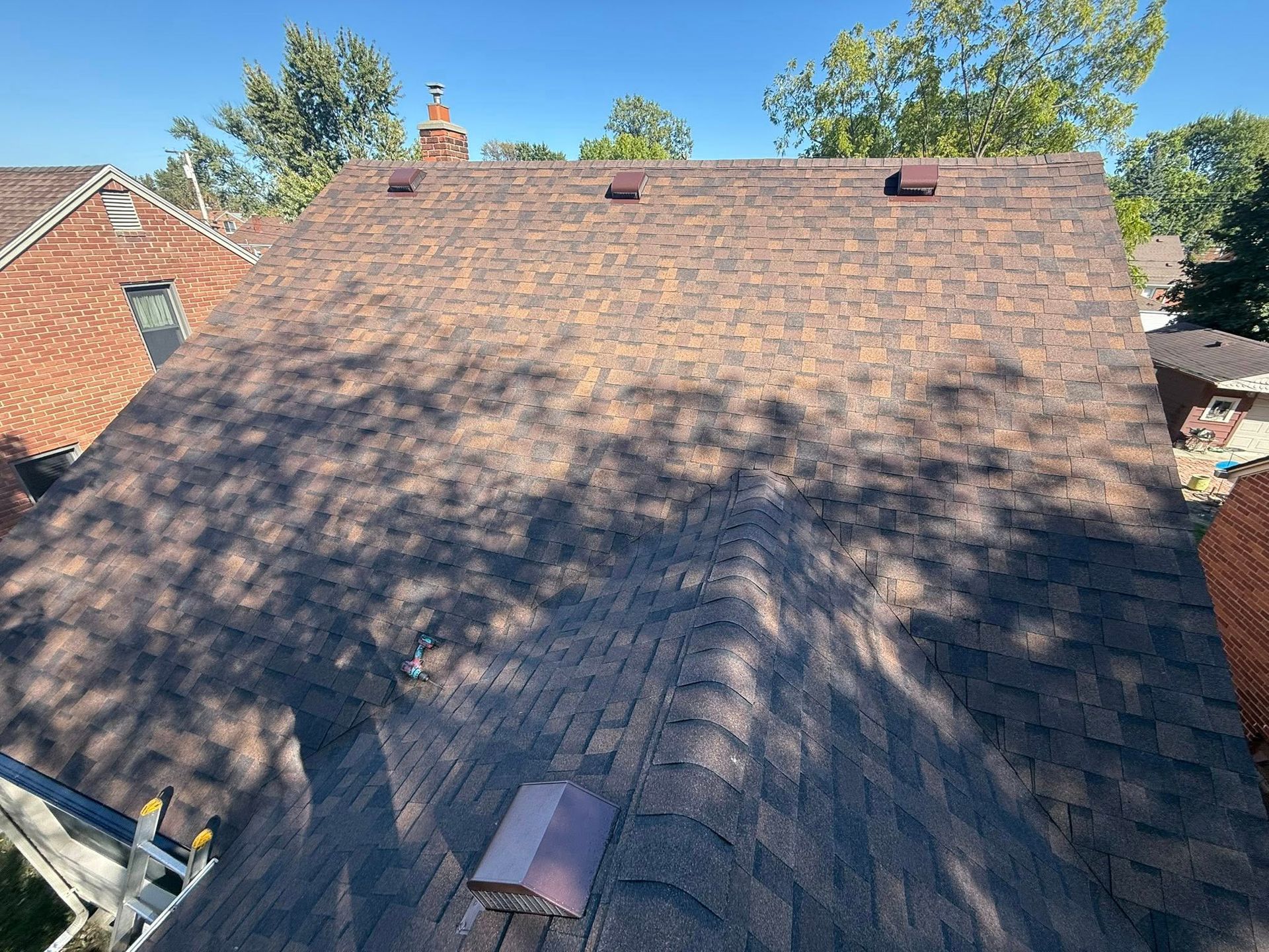 Brown shingled roof under a clear blue sky, partially shaded by tree branches.