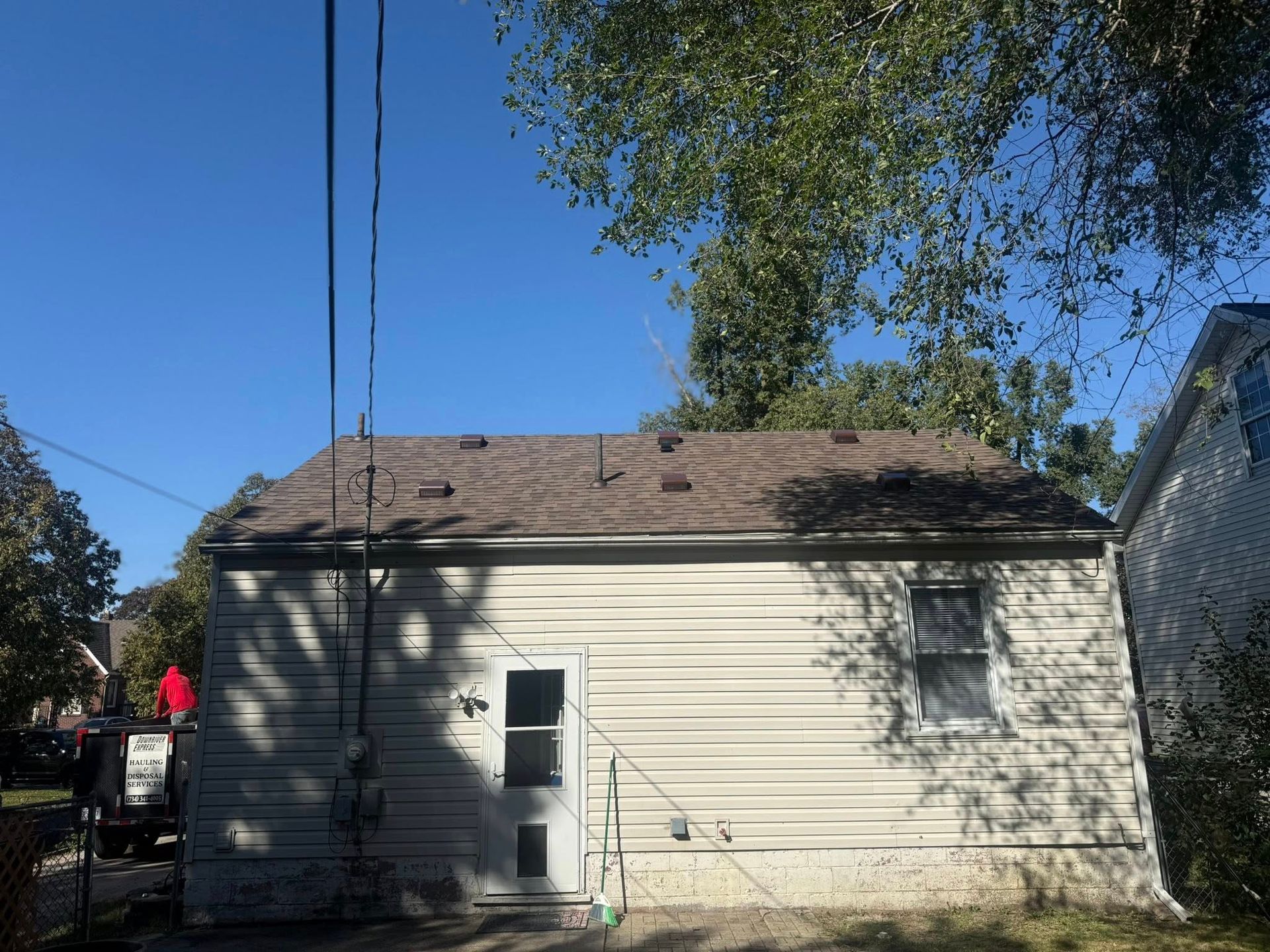 Back of a small, beige building with a brown roof and a white door, with utility lines overhead.