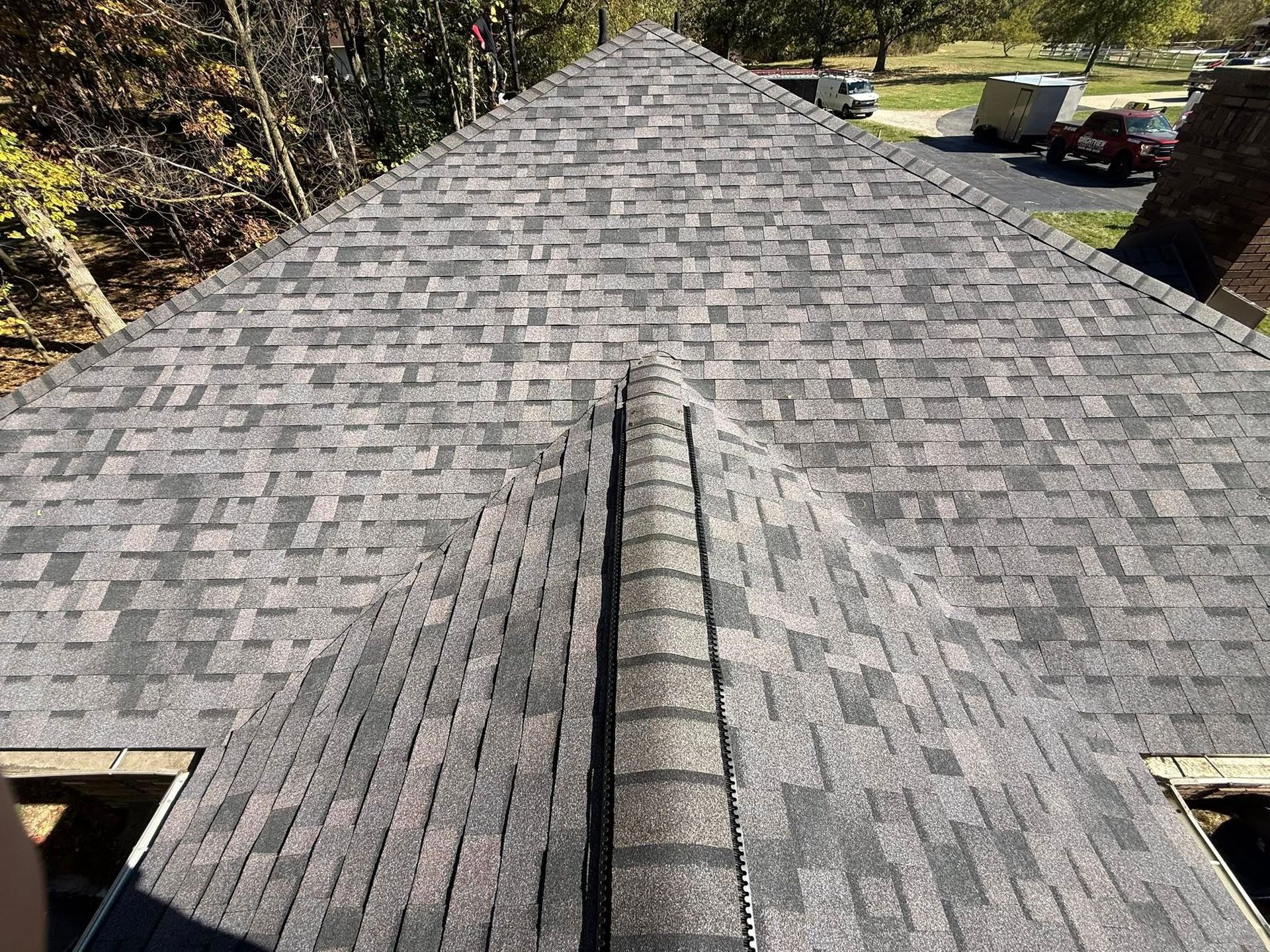 Close-up of a roof covered in gray and black shingles.  A central ridge is visible. Trees and vehicles in the distance.