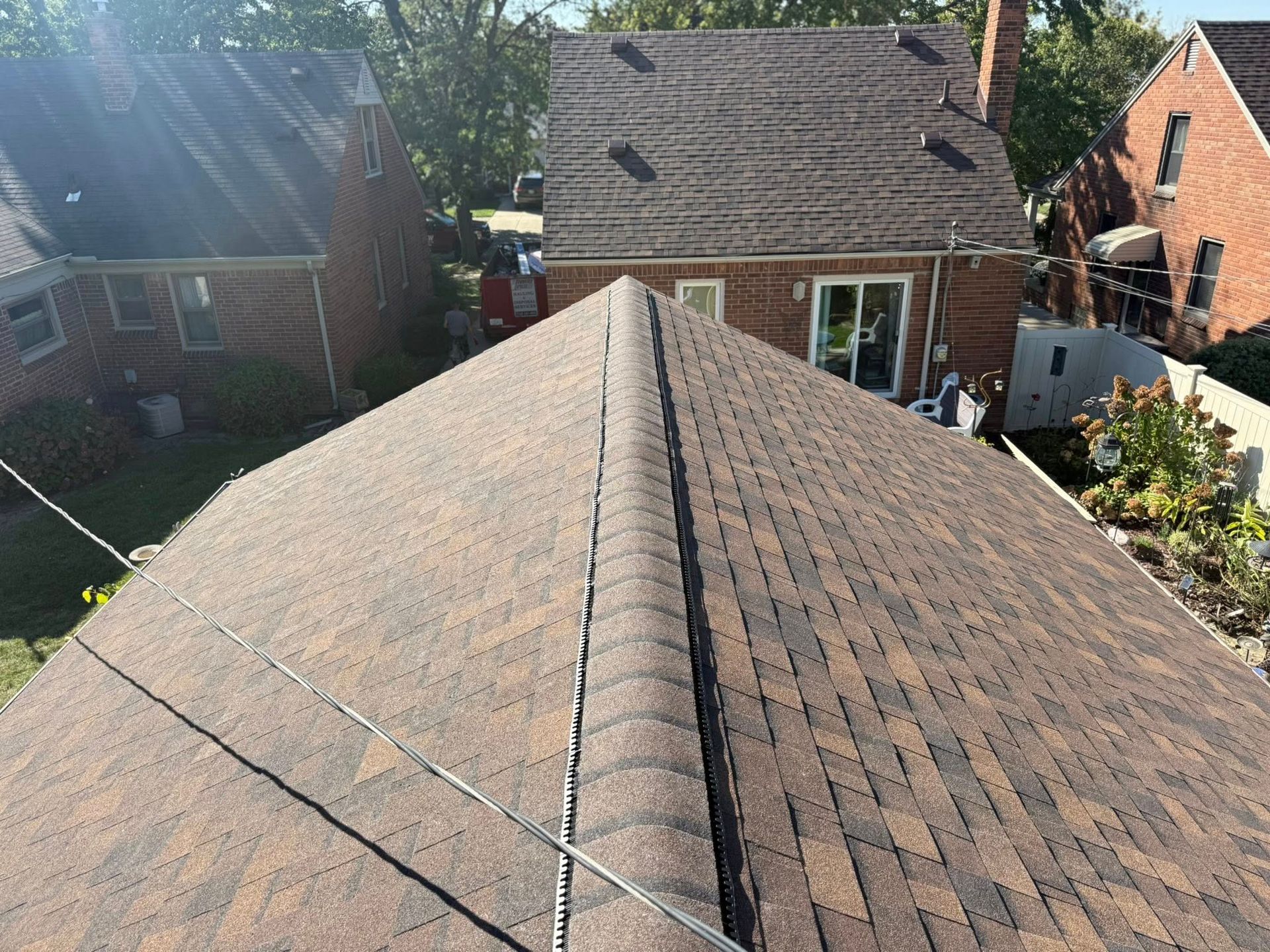 Brown shingle roof with a ridge, viewed from above, surrounded by other houses and trees on a sunny day.