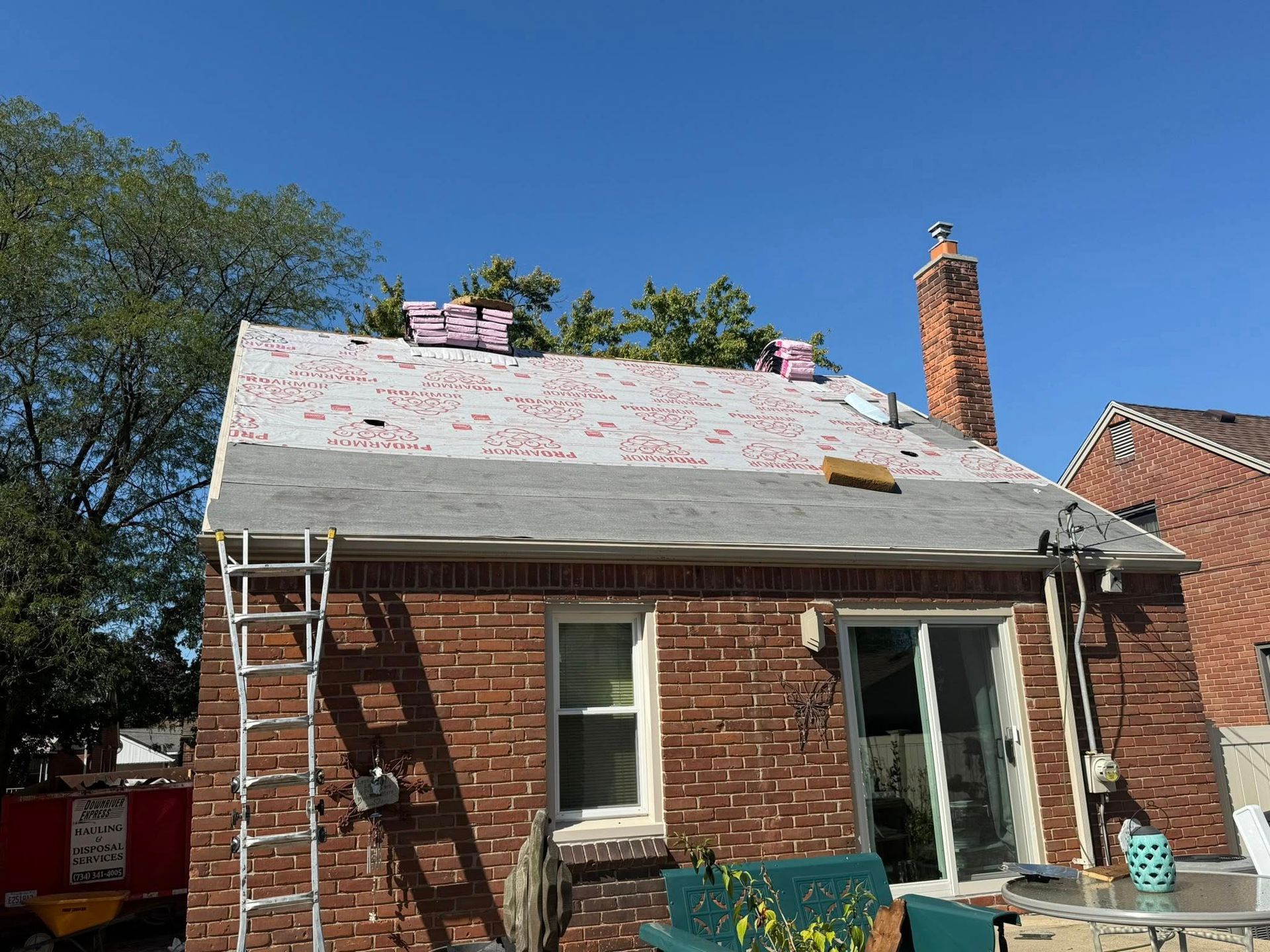 Roof under construction with shingles, on a brick house. Blue sky, ladder, and a chimney.